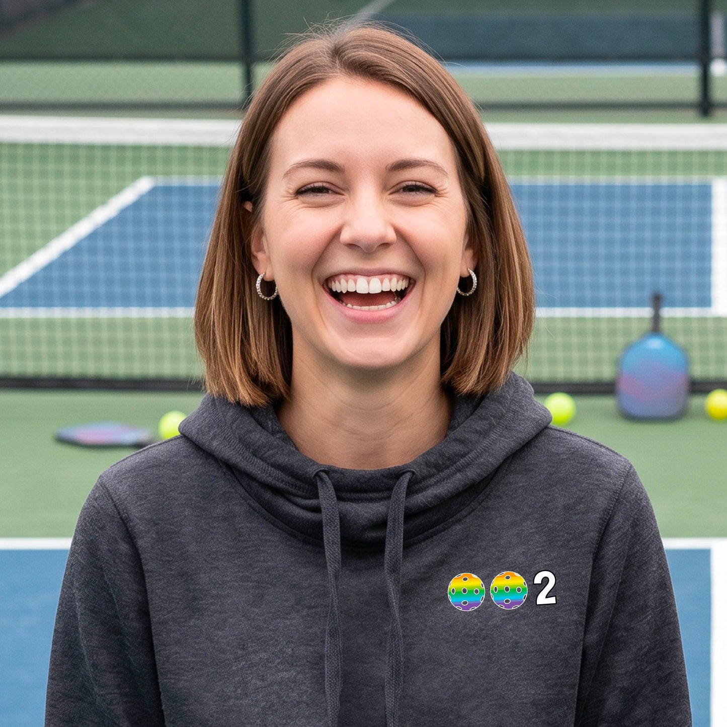 A woman smiling outdoors on a pickleball court, wearing a heasther navy blue hoodie with two pickleballs and the number 2 printed on the chest.
