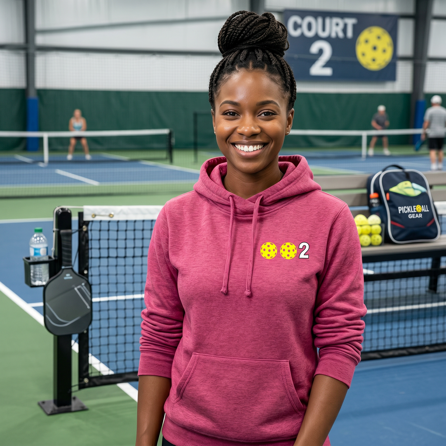 A woman smiling outdoors on a pickleball court, wearing a pink hoodie with two pickleballs and the number 2 printed on the chest.