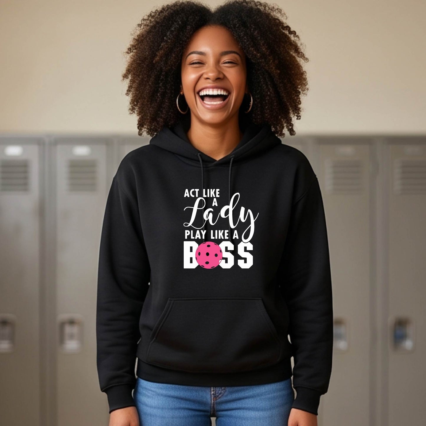 A woman wearing a black pullover hoodie with a white and pink pickleballl graphic text that reads 'ACT LIKE A Lady PLAY LIKE A BOSS' on the front, shown in a softly lit indoor setting.