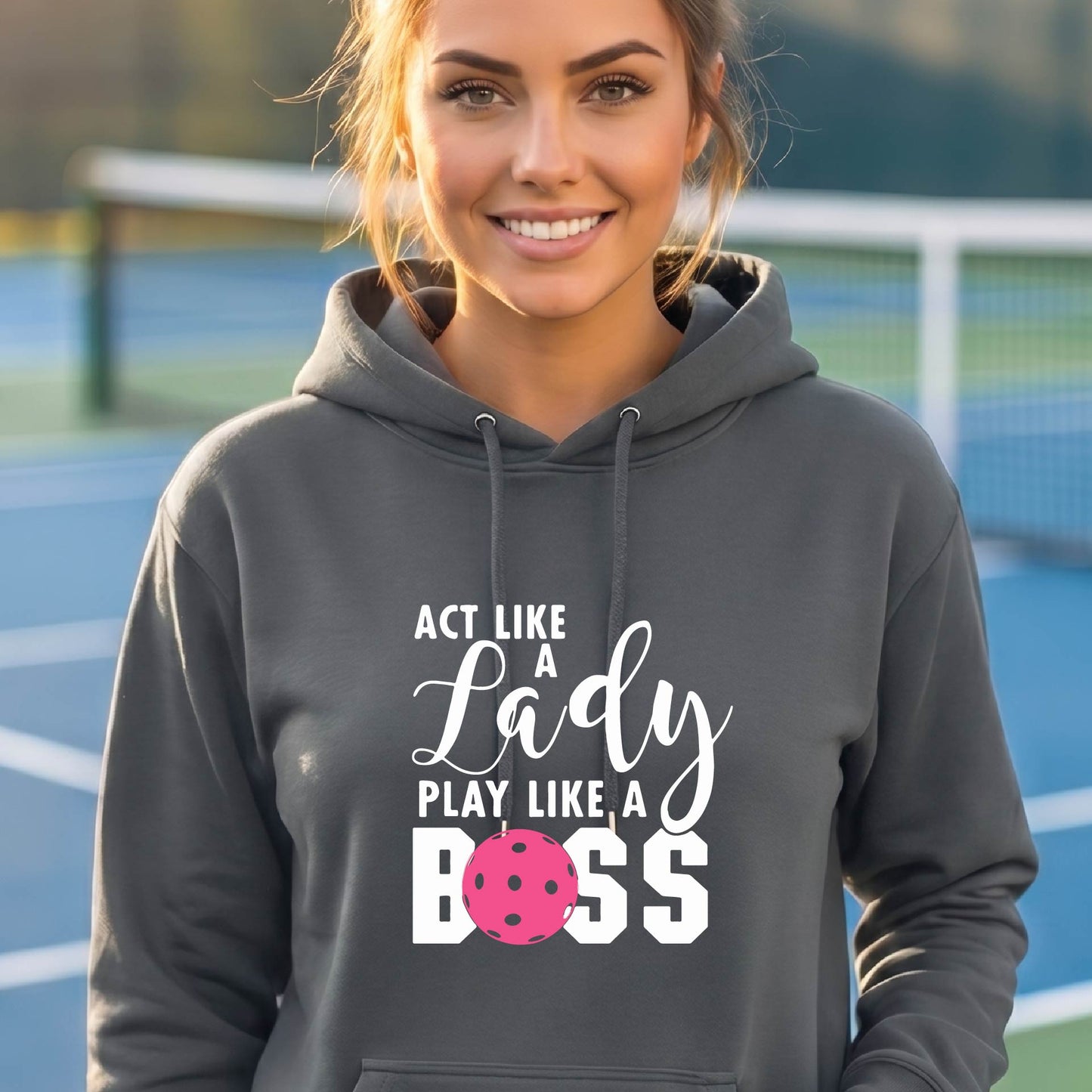 A woman wearing a gray pullover hoodie with a white and pink pickleballl graphic text that reads 'ACT LIKE A Lady PLAY LIKE A BOSS' on the front, shown in a softly lit indoor setting.