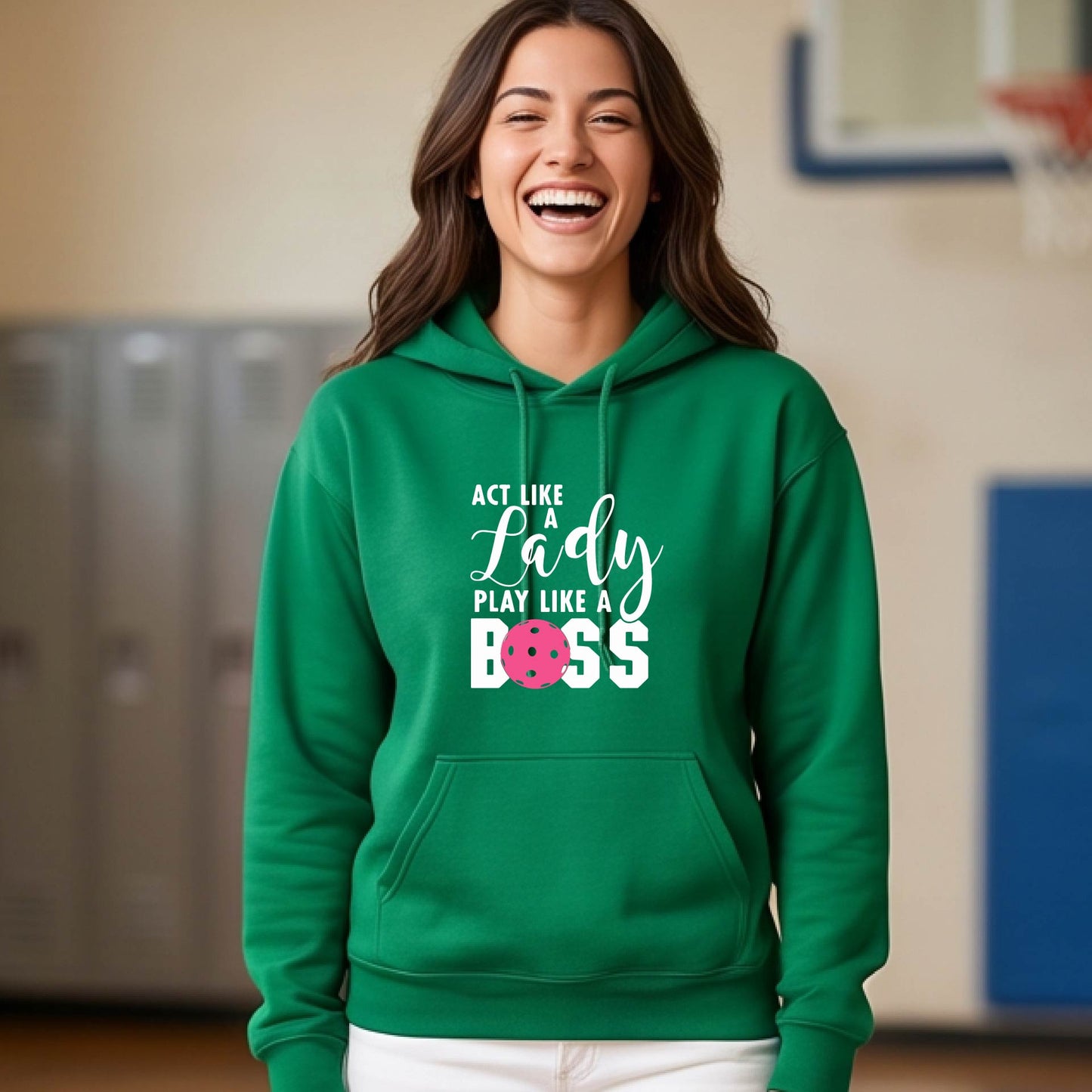 A woman wearing a green pullover hoodie with a white and pink pickleballl graphic text that reads 'ACT LIKE A Lady PLAY LIKE A BOSS' on the front, shown in a softly lit indoor setting.