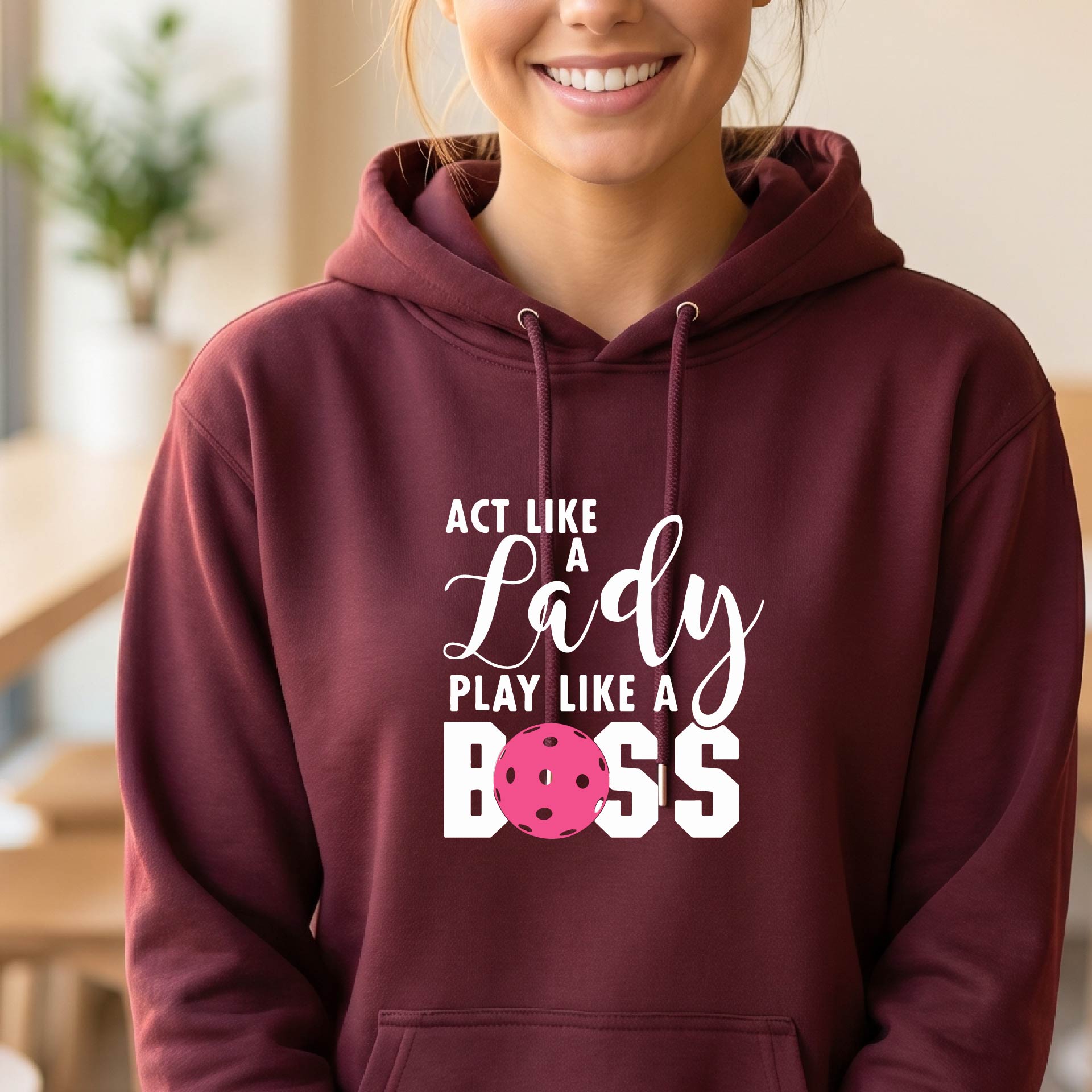 A woman wearing a maroon pullover hoodie with a white and pink pickleballl graphic text that reads 'ACT LIKE A Lady PLAY LIKE A BOSS' on the front, shown in a softly lit indoor setting.