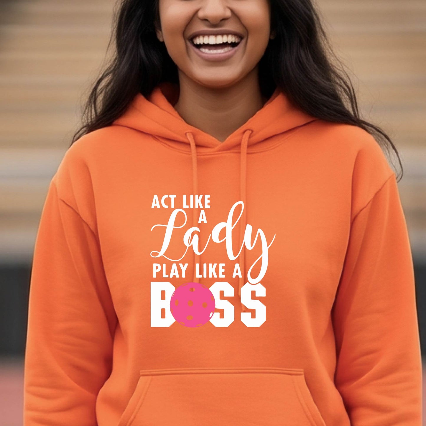 A woman wearing an orange pullover hoodie with a white and pink pickleballl graphic text that reads 'ACT LIKE A Lady PLAY LIKE A BOSS' on the front, shown in a softly lit indoor setting.