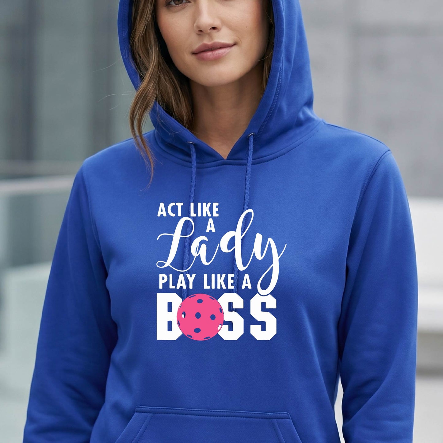 A woman wearing a royal blue pullover hoodie with a white and pink pickleballl graphic text that reads 'ACT LIKE A Lady PLAY LIKE A BOSS' on the front, shown in a softly lit indoor setting.