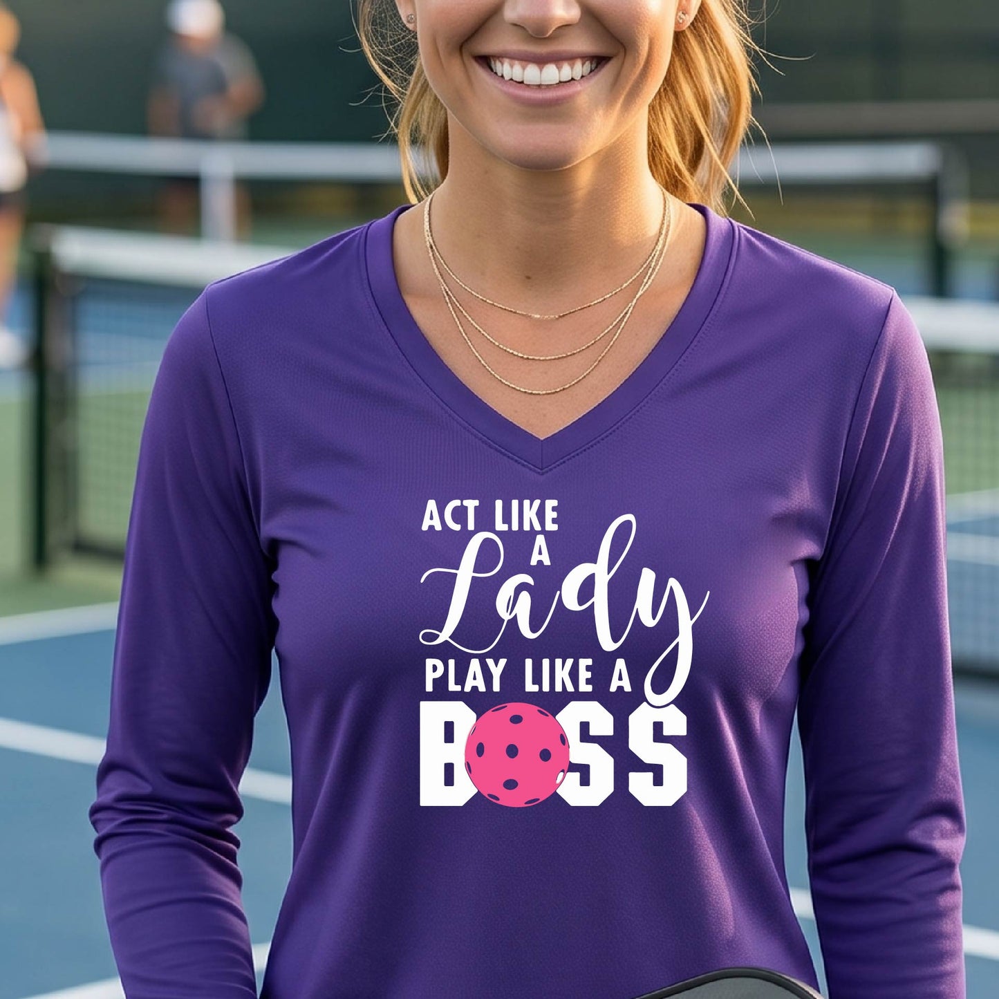 A woman on a pickleball court wearing a purple long-sleeve V-neck athletic shirt with the text 'ACT LIKE A Lady PLAY LIKE A BOSS' and a pink pickleball graphic.