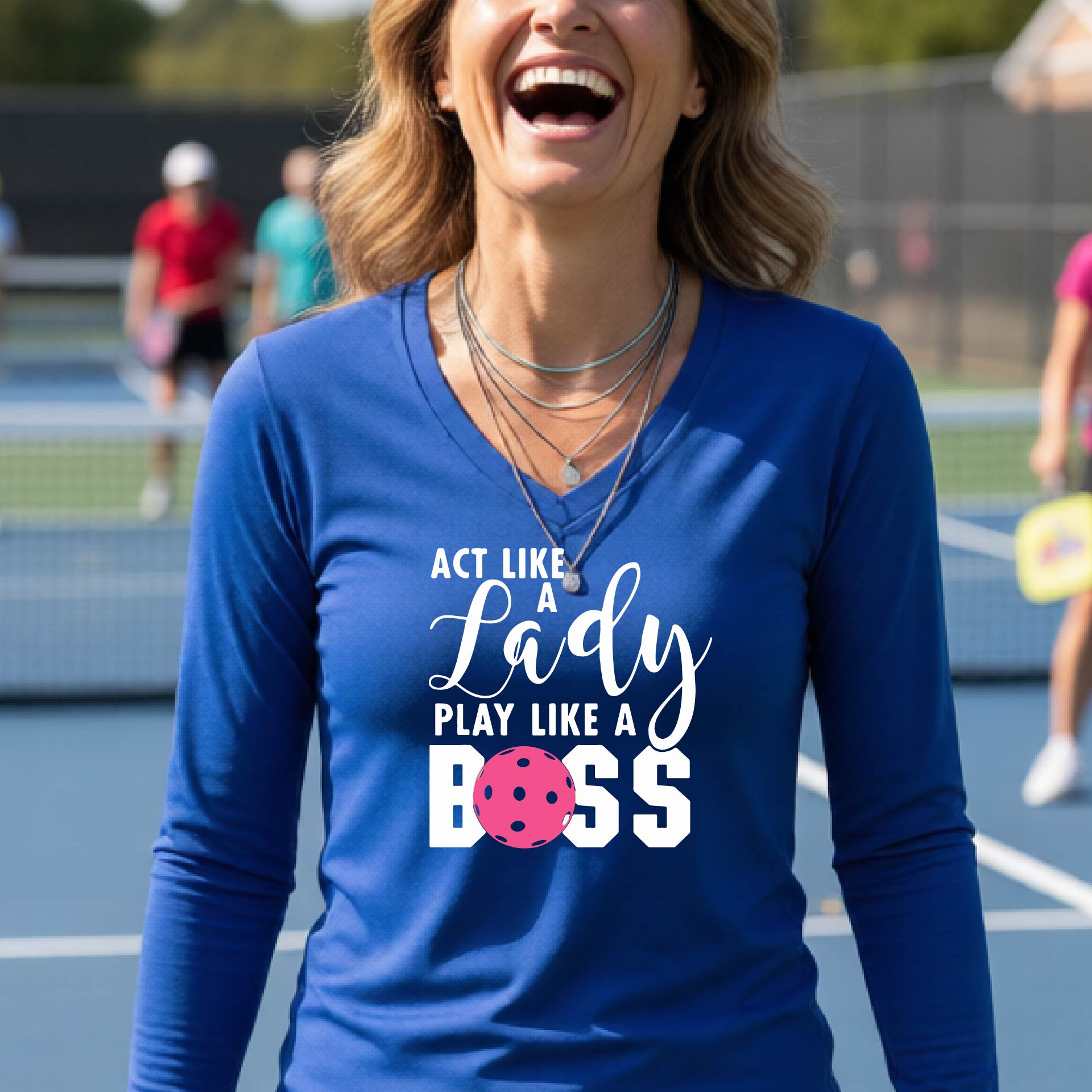 A woman on a pickleball court wearing a royal blue long-sleeve V-neck athletic shirt with the text 'ACT LIKE A Lady PLAY LIKE A BOSS' and a pink pickleball graphic.