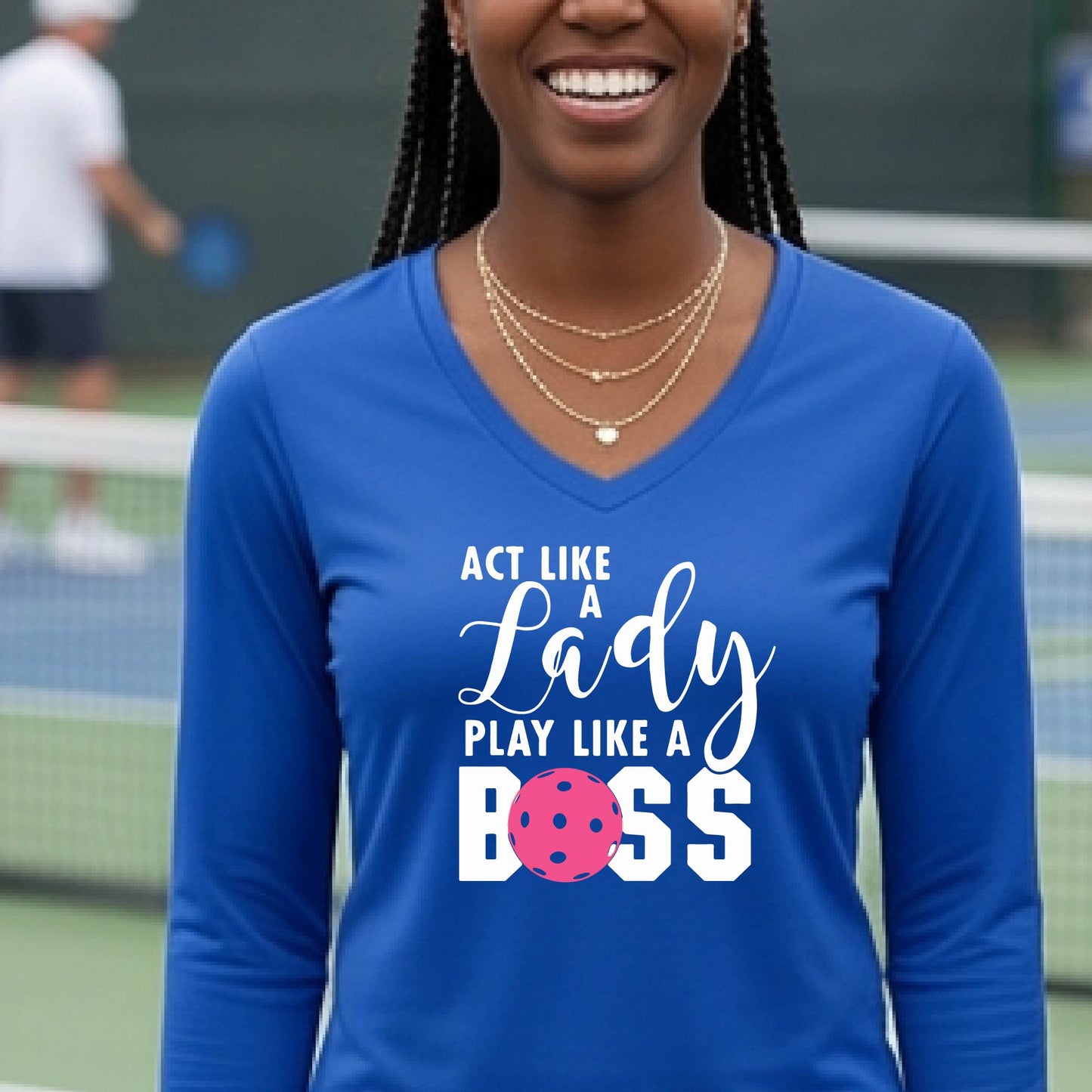 A woman on a pickleball court wearing a bright royal blue long-sleeve V-neck shirt with the text 'ACT LIKE A Lady PLAY LIKE A BOSS' in white, with a pink pickleball graphic replacing the letter O in BOSS.