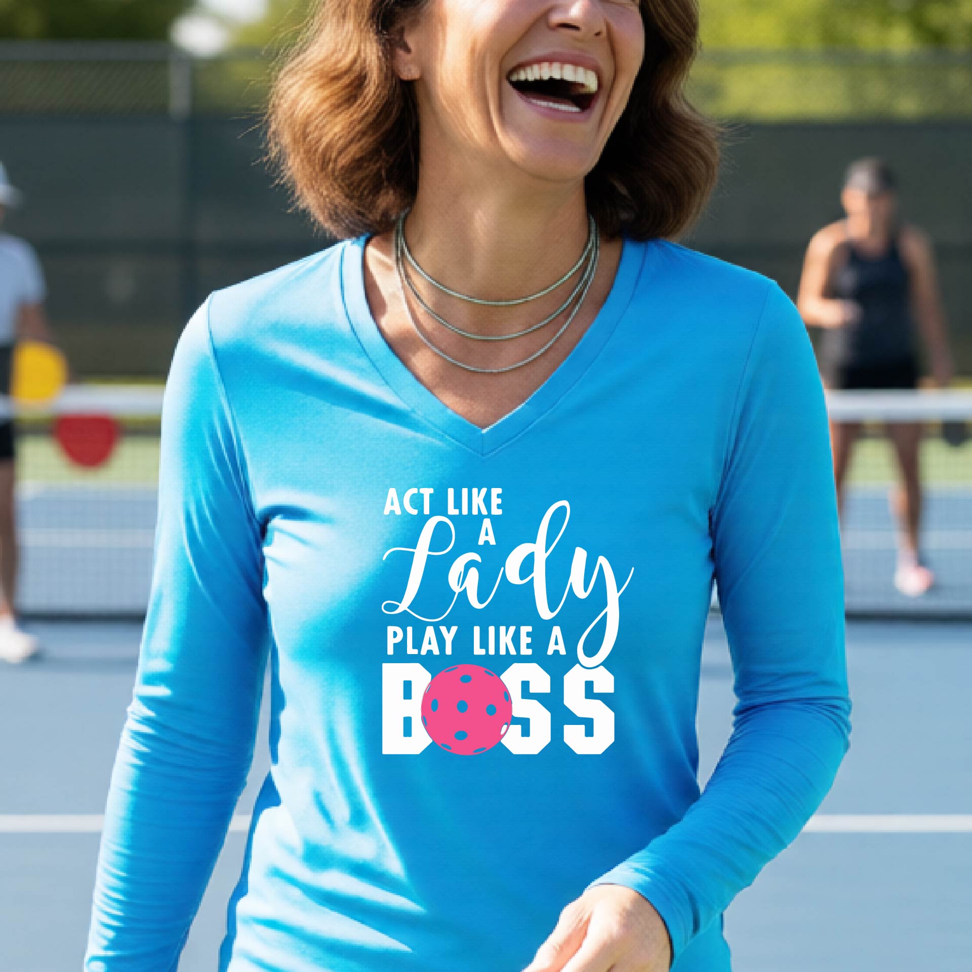 A woman on a pickleball court wearing a bright blue long-sleeve V-neck shirt with the text 'ACT LIKE A Lady PLAY LIKE A BOSS' in white, with a pink pickleball graphic replacing the letter O in BOSS.