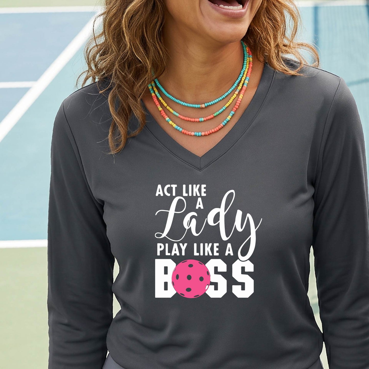 A woman on a pickleball court wearing a dark gray long-sleeve V-neck athletic shirt with the text 'ACT LIKE A Lady PLAY LIKE A BOSS' and a pink pickleball graphic.
