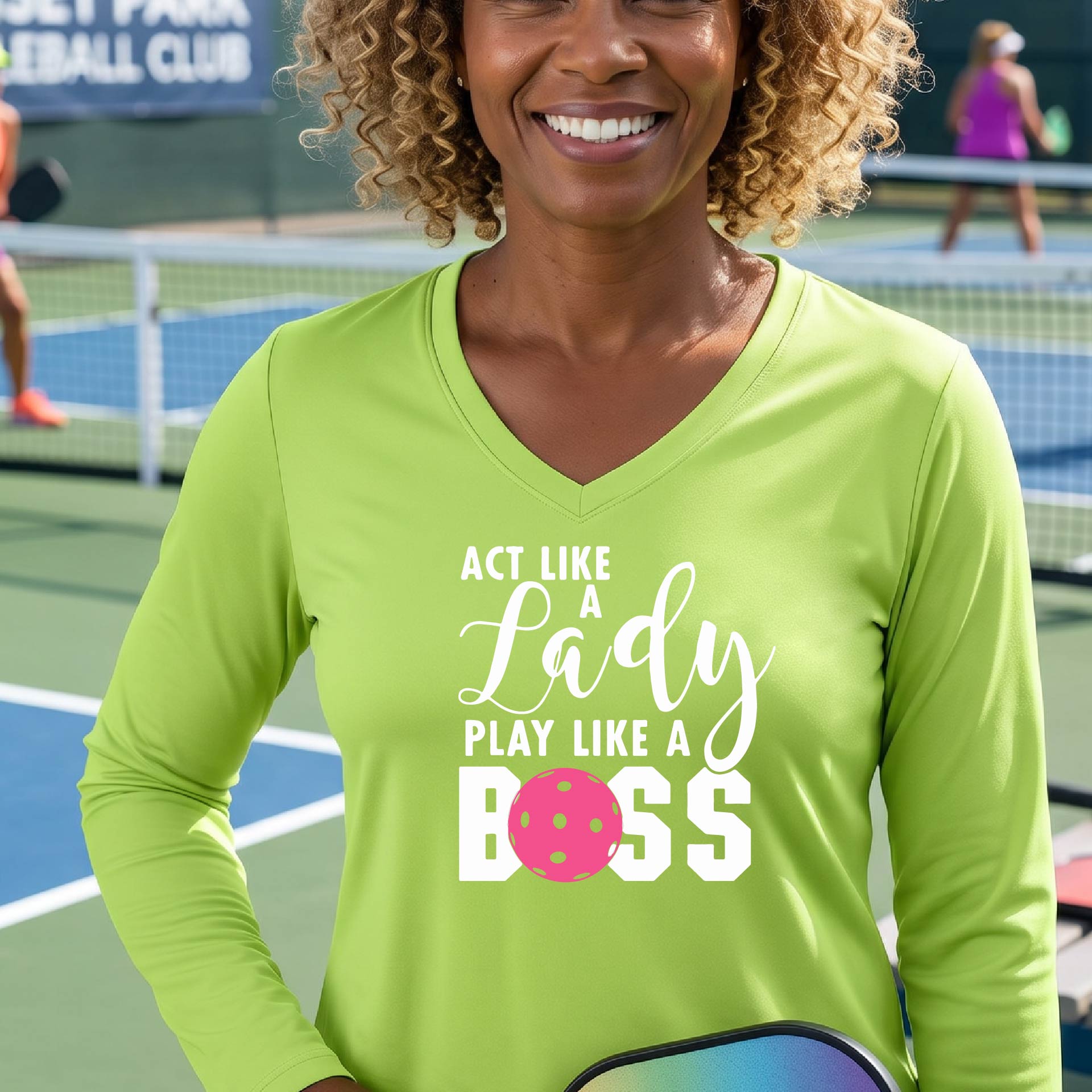 A woman on a pickleball court wearing a bright lime green long-sleeve V-neck athletic shirt with the text 'ACT LIKE A Lady PLAY LIKE A BOSS' and a pink pickleball graphic.