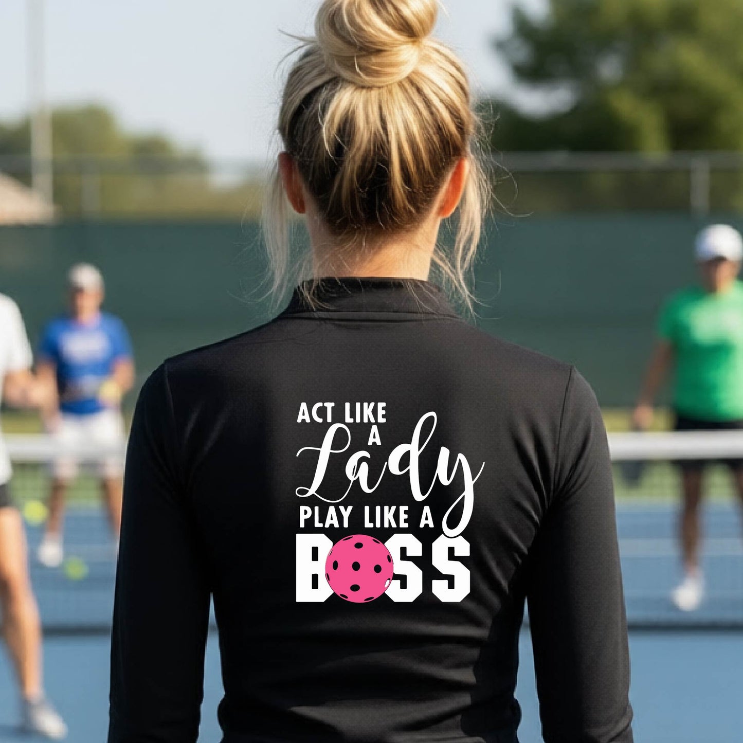 A woman wearing a black quarter-zip long-sleeve athletic shirt on a pickleball court. The back of the shirt features the text 'ACT LIKE A Lady PLAY LIKE A BOSS' in white, with a pink pickleball graphic replacing the letter 'O' in 'BOSS'.