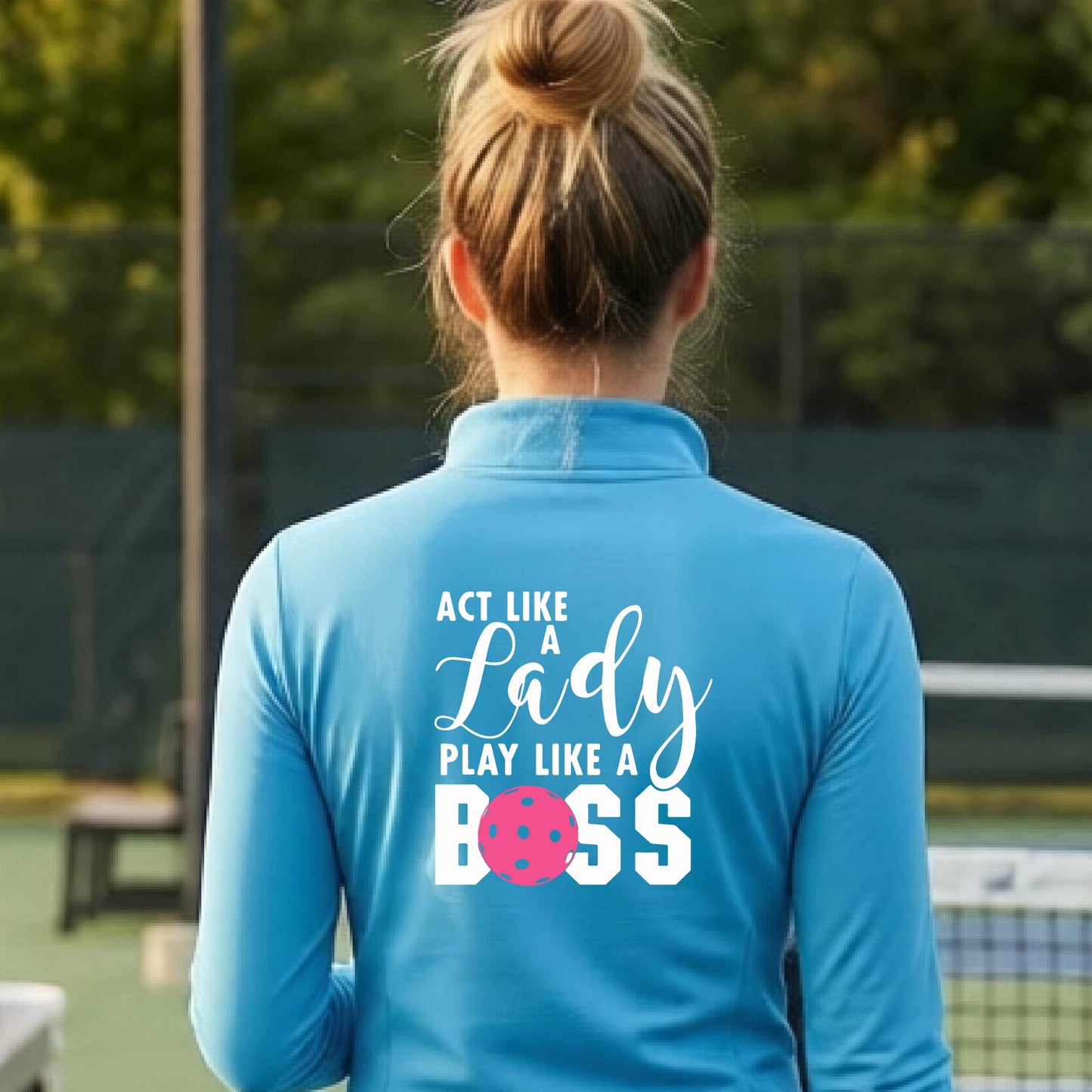 A woman wearing a light blue quarter-zip long-sleeve athletic shirt on a pickleball court. The back of the shirt features the text 'ACT LIKE A Lady PLAY LIKE A BOSS' in white, with a pink pickleball graphic replacing the letter 'O' in 'BOSS'.