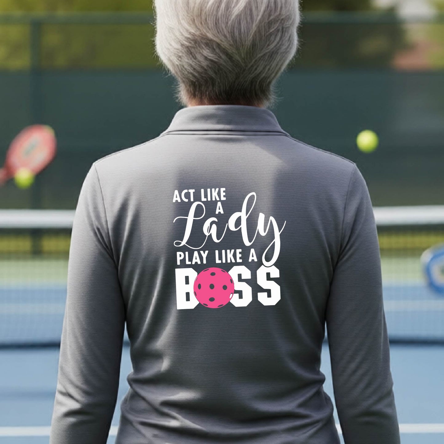 A woman wearing a gray quarter-zip long-sleeve athletic shirt on a pickleball court. The back of the shirt features the text 'ACT LIKE A Lady PLAY LIKE A BOSS' in white, with a pink pickleball graphic replacing the letter 'O' in 'BOSS'.