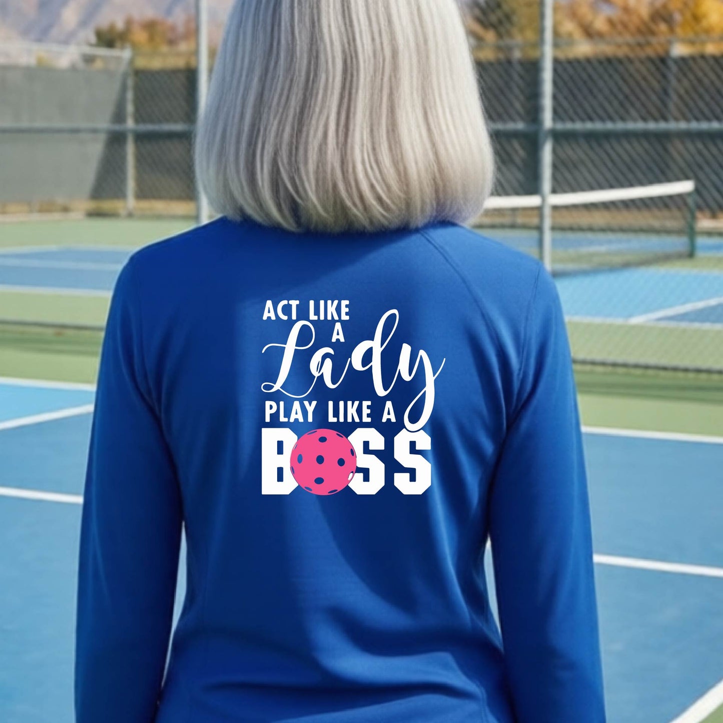 A woman wearing a royal blue quarter-zip long-sleeve athletic shirt on a pickleball court. The back of the shirt features the text 'ACT LIKE A Lady PLAY LIKE A BOSS' in white, with a pink pickleball graphic replacing the letter 'O' in 'BOSS'.