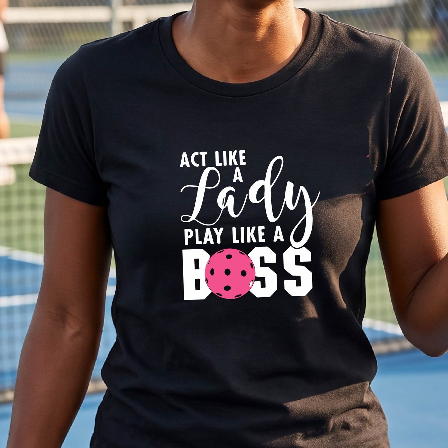 A woman on a pickleball court wearing a black graphic t-shirt that reads 'ACT LIKE A Lady PLAY LIKE A BOSS' with a pink pickleball graphic, holding a paddle.