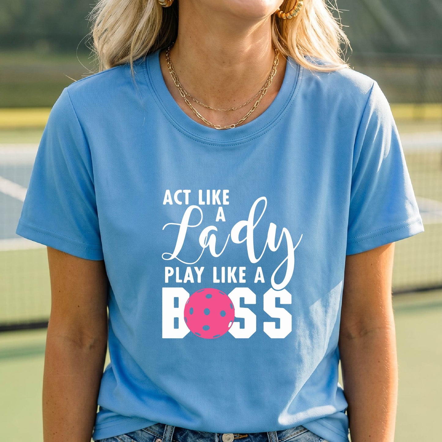 A woman on a pickleball court wearing a blue graphic t-shirt that reads 'ACT LIKE A Lady PLAY LIKE A BOSS' with a pink pickleball graphic, holding a paddle.