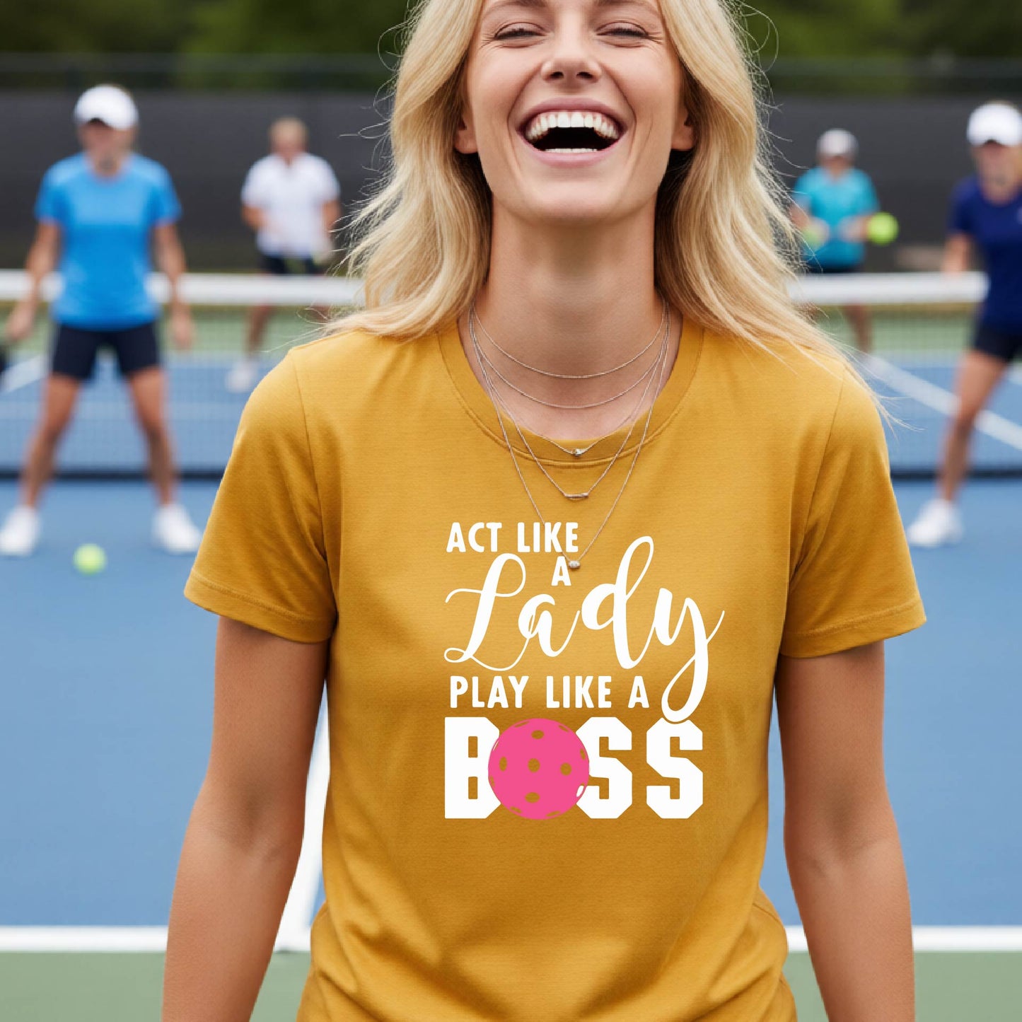 A woman on a pickleball court wearing a gold graphic t-shirt that reads 'ACT LIKE A Lady PLAY LIKE A BOSS' with a pink pickleball graphic, holding a paddle.