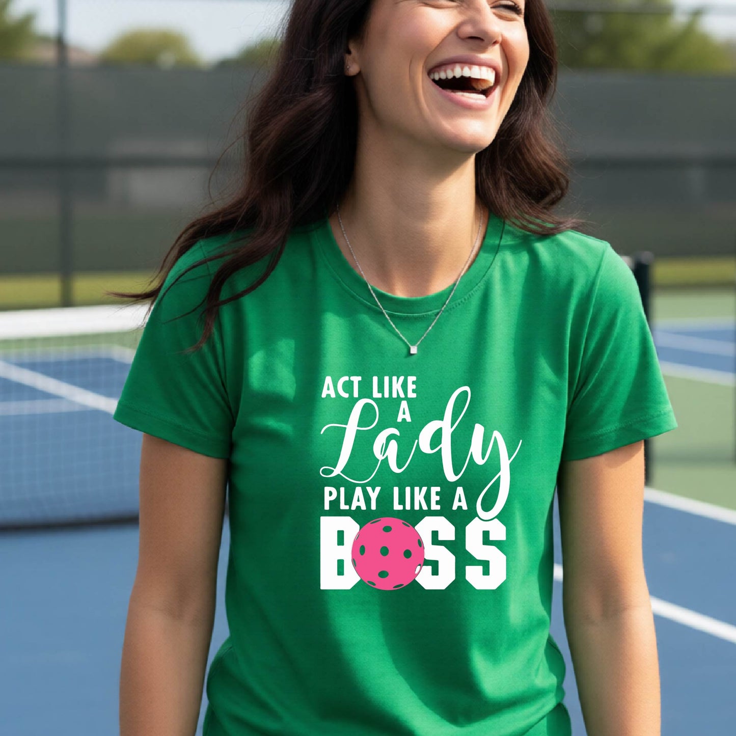 A woman on a pickleball court wearing a green graphic t-shirt that reads 'ACT LIKE A Lady PLAY LIKE A BOSS' with a pink pickleball graphic, holding a paddle.