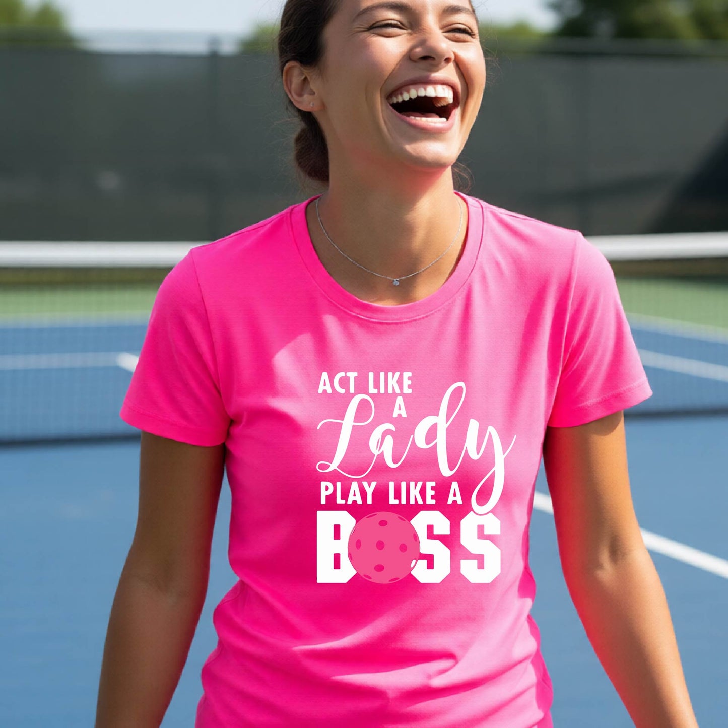 A woman on a pickleball court wearing a pink graphic t-shirt that reads 'ACT LIKE A Lady PLAY LIKE A BOSS' with a pink pickleball graphic, holding a paddle.