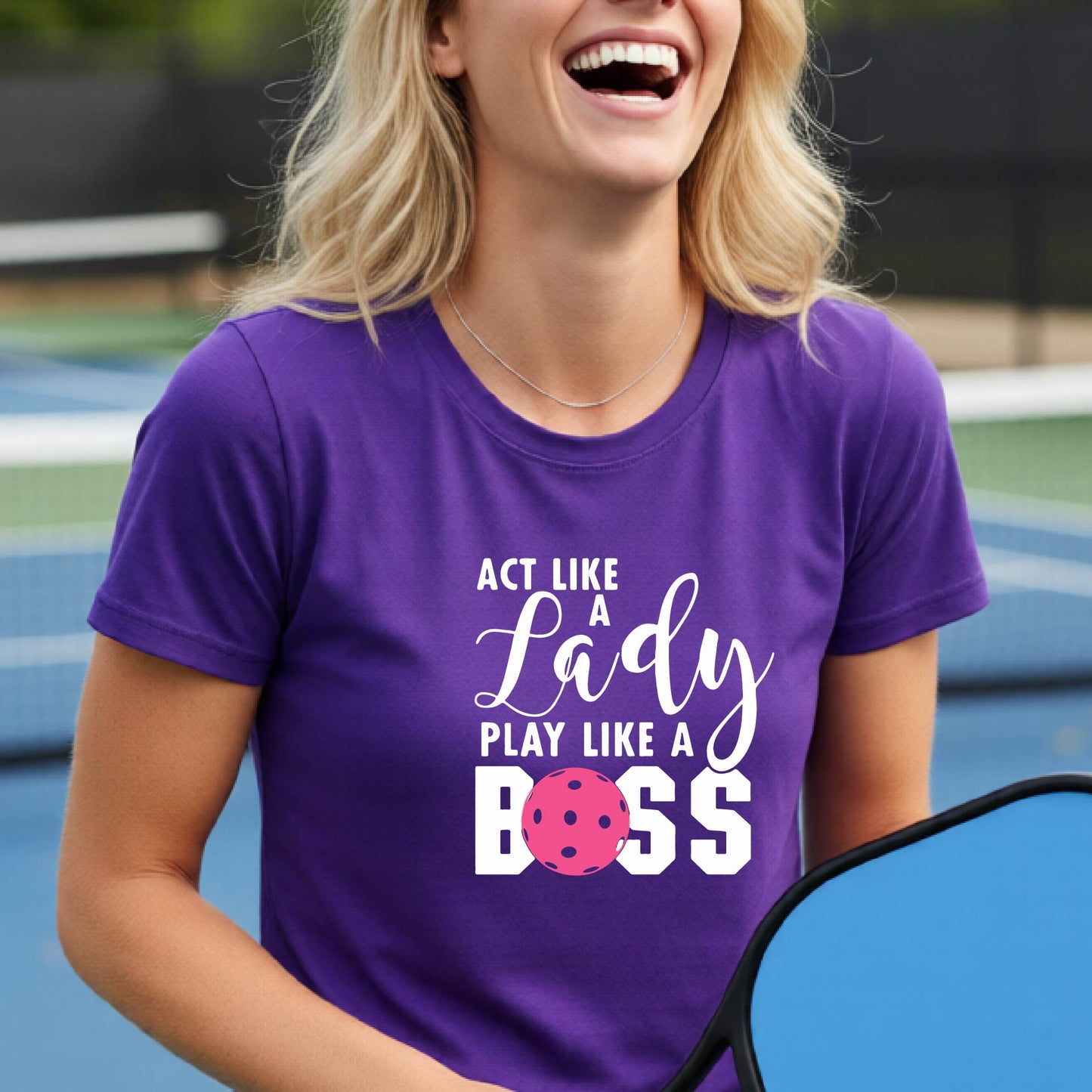 A woman on a pickleball court wearing a purple graphic t-shirt that reads 'ACT LIKE A Lady PLAY LIKE A BOSS' with a pink pickleball graphic, holding a paddle.