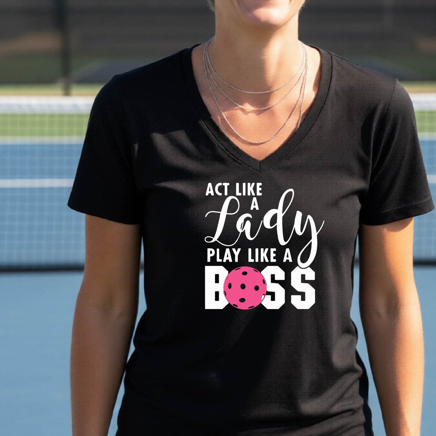 A woman wearing a black V-neck t-shirt with a graphic print that reads 'ACT LIKE A Lady PLAY LIKE A BOSS' in white, featuring a pink pickleall ball graphic for the letter 'O'. 