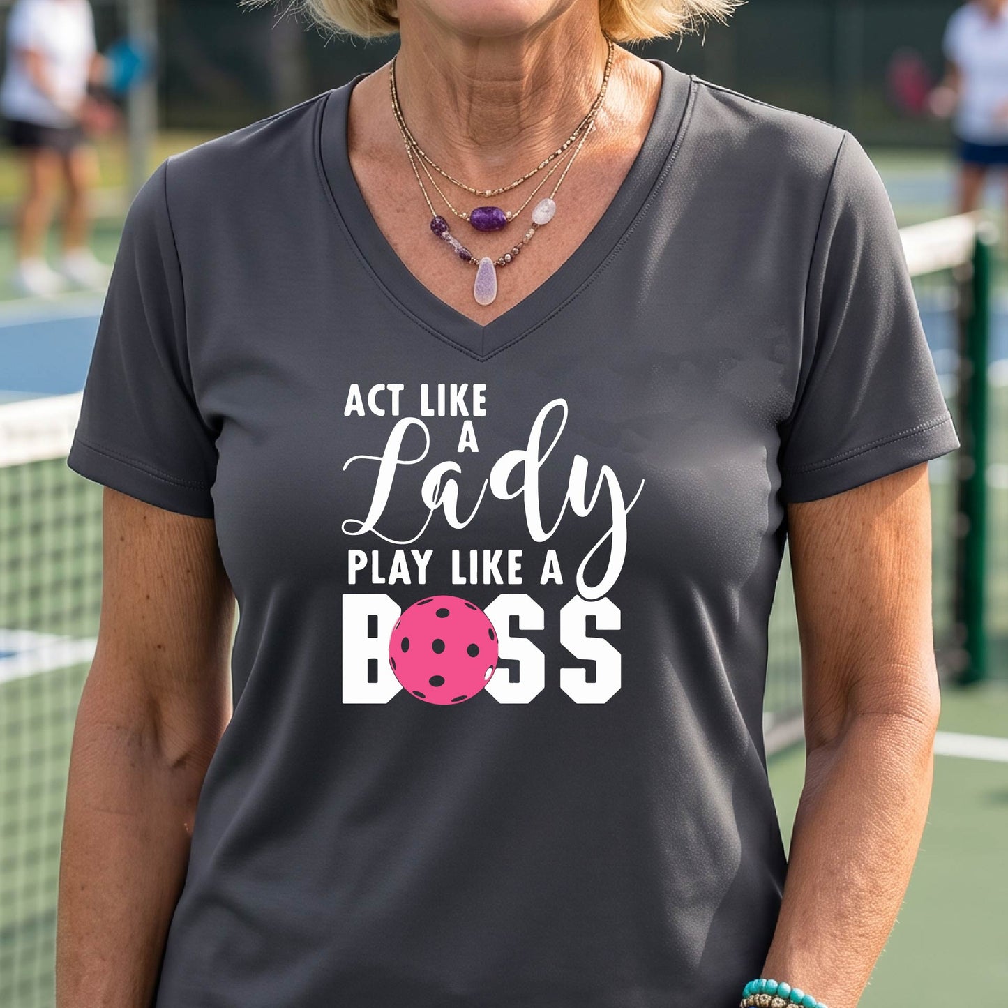 A woman wearing a dark grey V-neck t-shirt with a graphic print that reads 'ACT LIKE A Lady PLAY LIKE A BOSS' in white, featuring a pink pickleall ball graphic for the letter 'O'. 