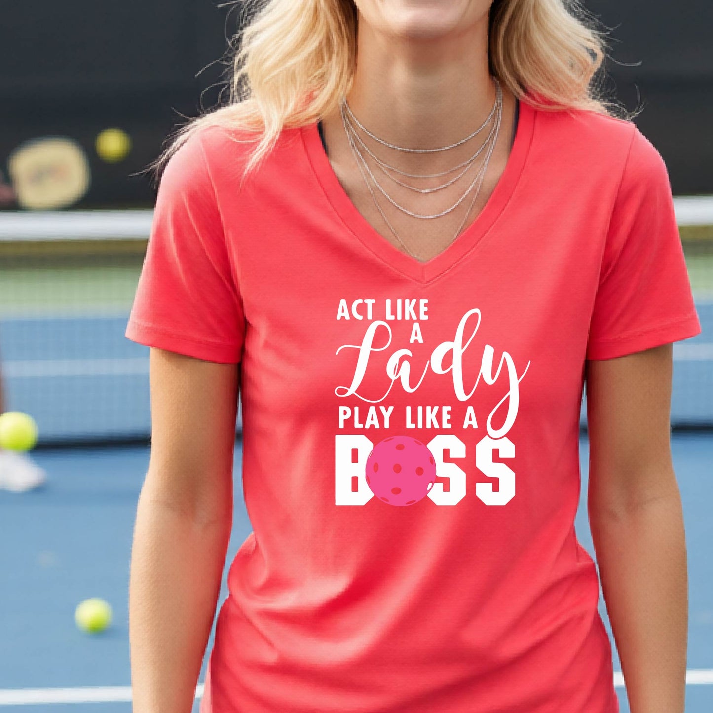 A woman wearing a coral pink V-neck t-shirt with a graphic print that reads 'ACT LIKE A Lady PLAY LIKE A BOSS' in white, featuring a pink pickleall ball graphic for the letter 'O'. 