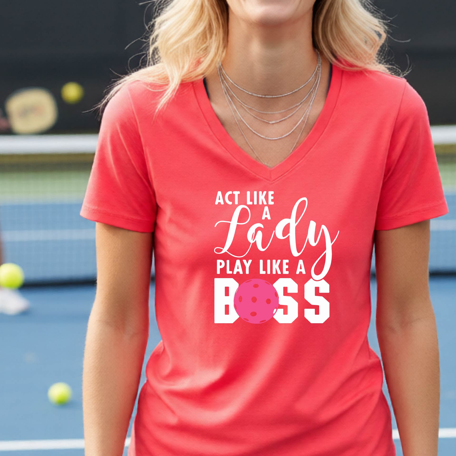 A woman wearing a coral pink V-neck t-shirt with a graphic print that reads 'ACT LIKE A Lady PLAY LIKE A BOSS' in white, featuring a pink pickleall ball graphic for the letter 'O'. 