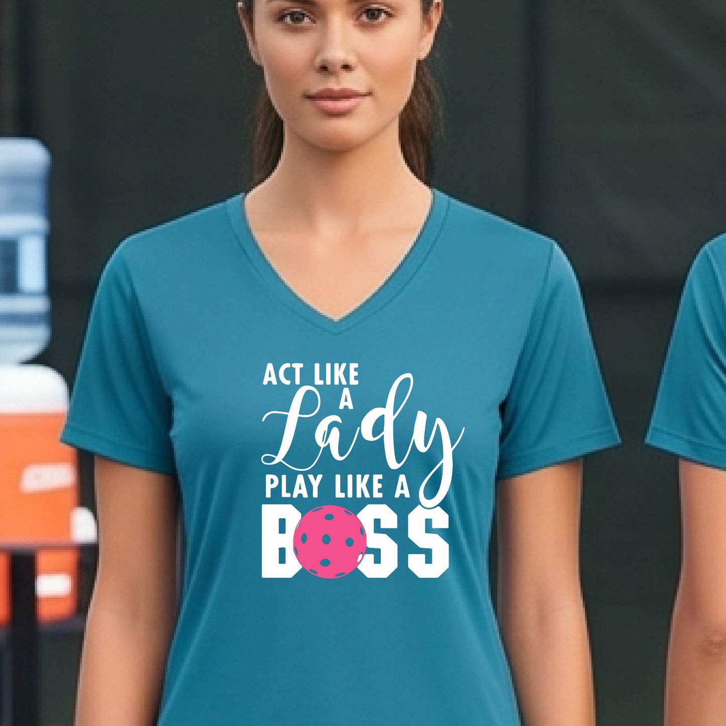 A woman wearing a teal V-neck t-shirt with a graphic print that reads 'ACT LIKE A Lady PLAY LIKE A BOSS' in white, featuring a pink pickleall ball graphic for the letter 'O'. 
