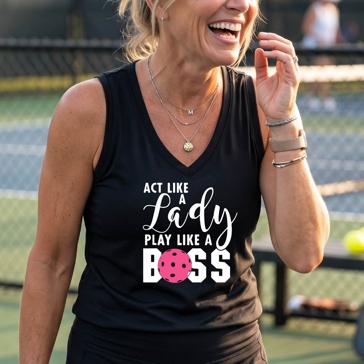 A woman on a pickkleball court wearing a black sleeveless tank top with a white and pink graphic that reads 'ACT LIKE A Lady PLAY LIKE A BOSS'.