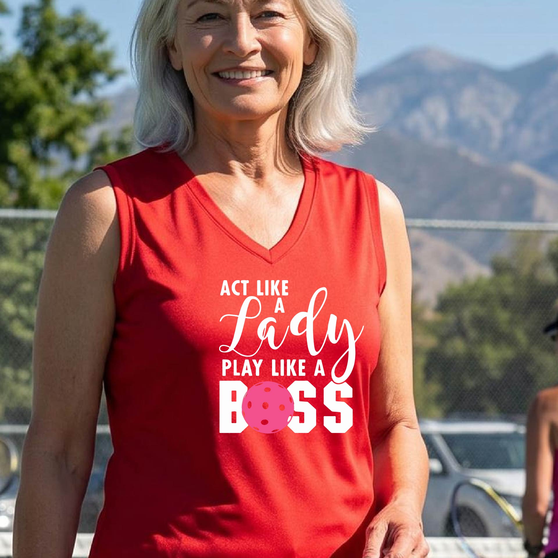 A woman on a pickkleball court wearing a red sleeveless tank top with a white and pink graphic that reads 'ACT LIKE A Lady PLAY LIKE A BOSS'.