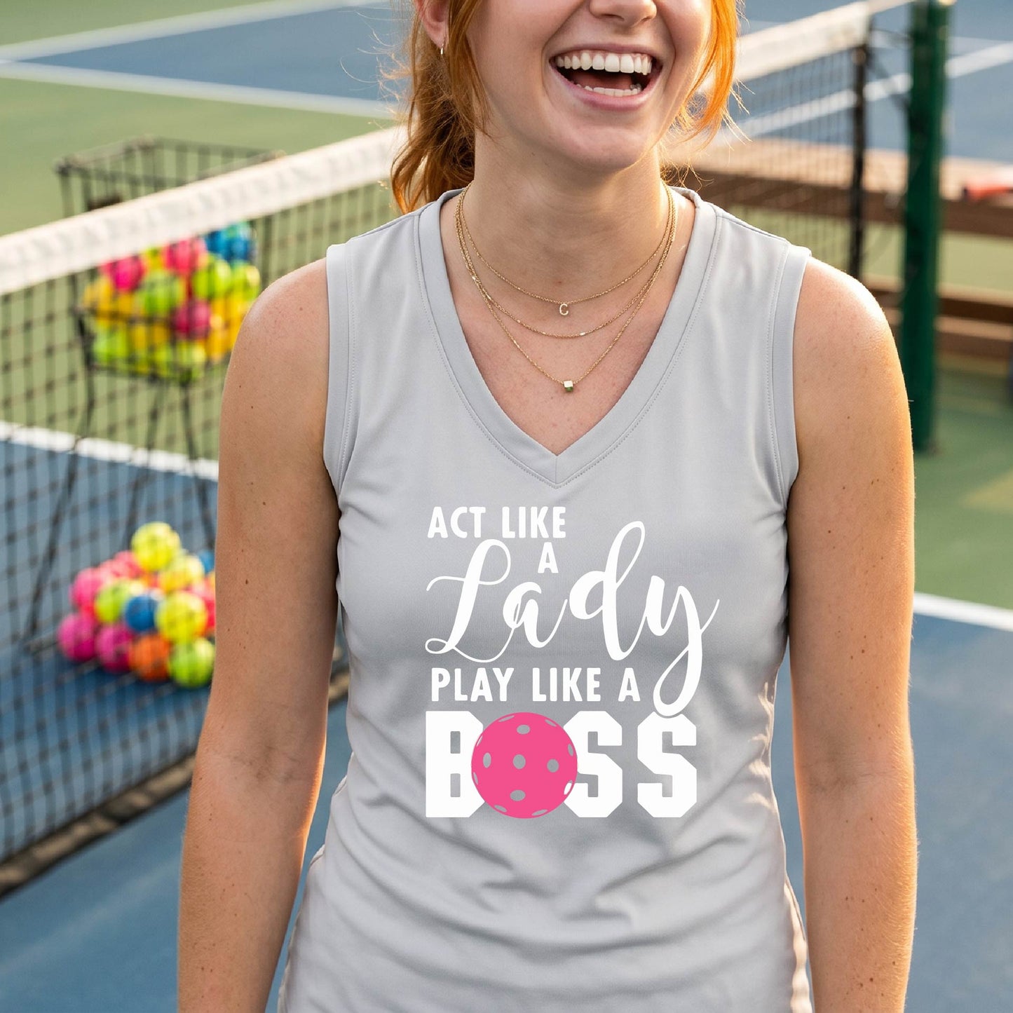 A woman on a pickkleball court wearing a light gray sleeveless tank top with a white and pink graphic that reads 'ACT LIKE A Lady PLAY LIKE A BOSS'.