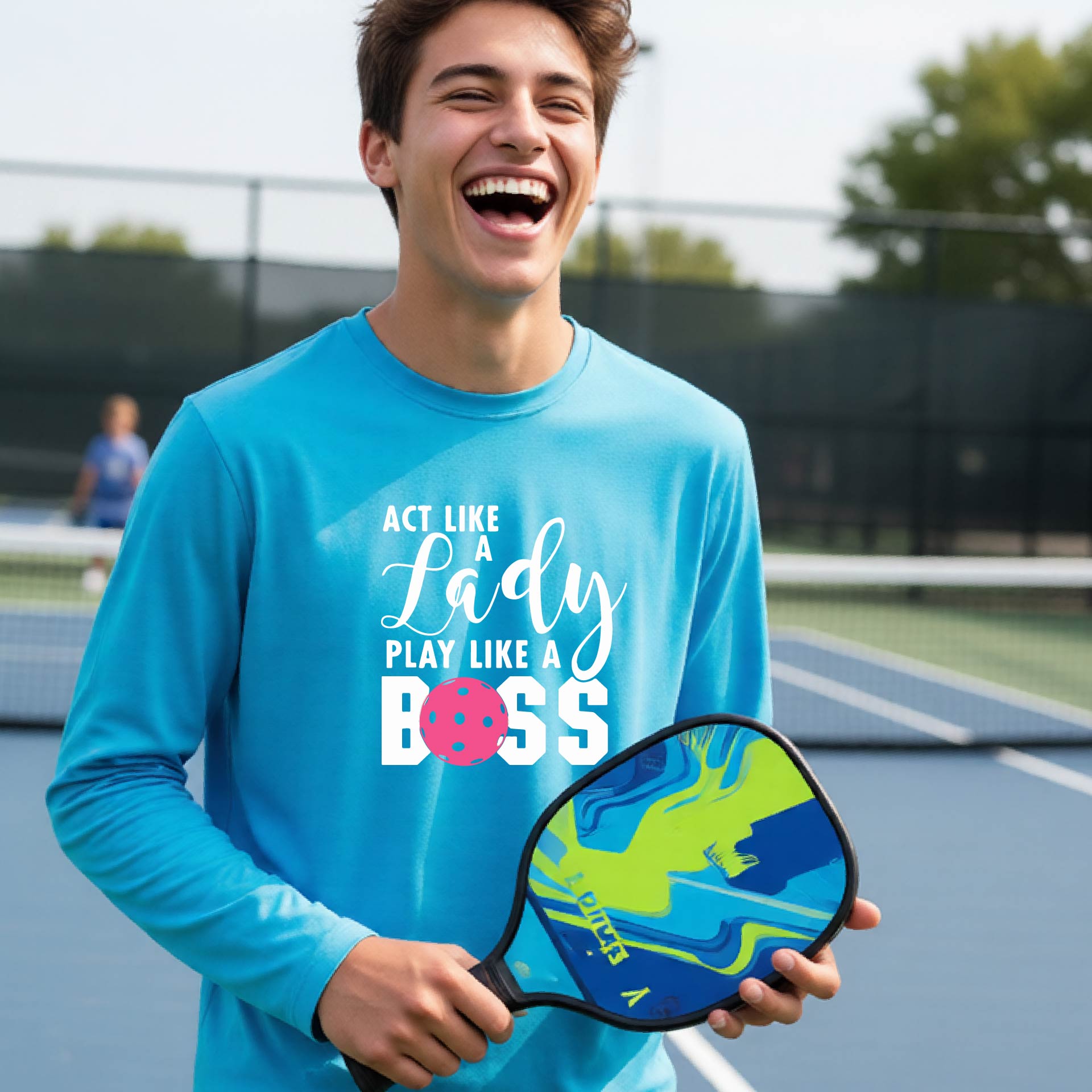 A young person laughing on a pickleball court wearing a light blue long-sleeve t-shirt with the text 'ACT LIKE A Lady PLAY LIKE A BOSS' and a pink polka-dot graphic.