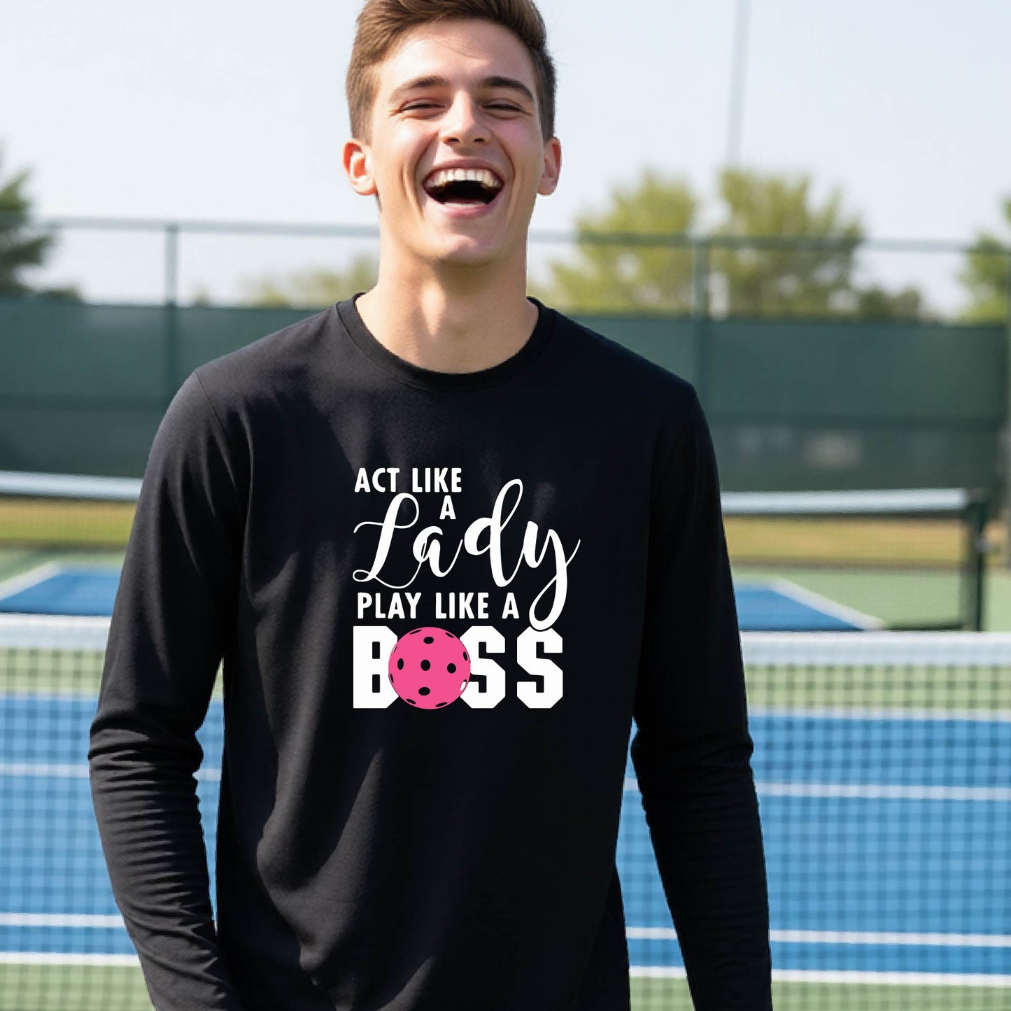 A young person laughing on a pickleball court wearing a black long-sleeve t-shirt with the text 'ACT LIKE A Lady PLAY LIKE A BOSS' and a pink polka-dot graphic.
