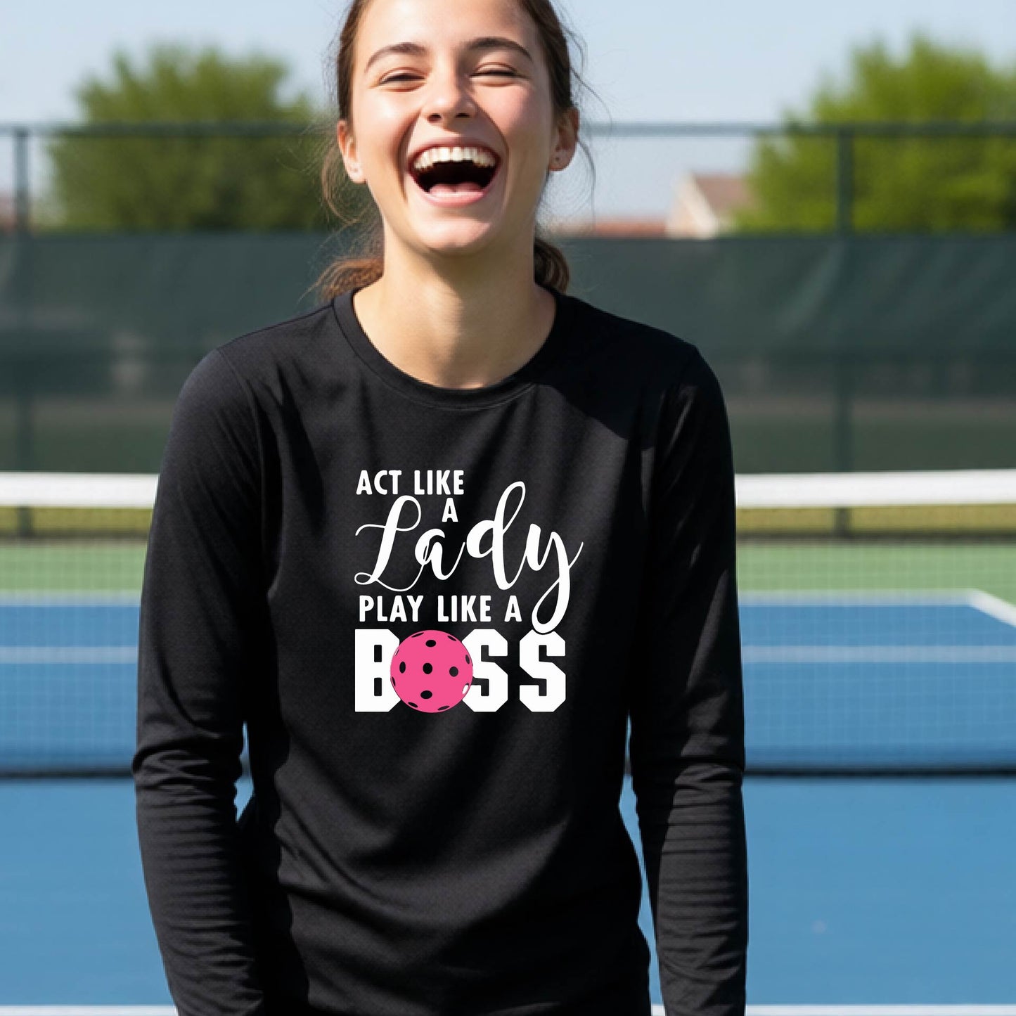 A young person laughing on a pickleball court wearing a black long-sleeve t-shirt with the text 'ACT LIKE A Lady PLAY LIKE A BOSS' and a pink polka-dot graphic.