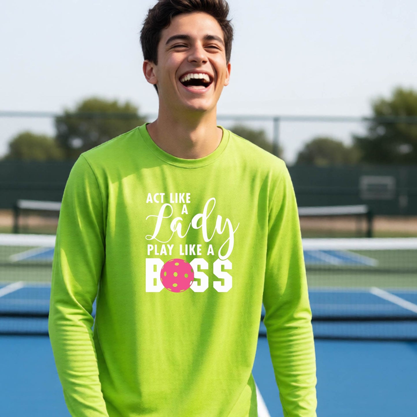 A young person laughing on a pickleball court wearing a lime green long-sleeve t-shirt with the text 'ACT LIKE A Lady PLAY LIKE A BOSS' and a pink polka-dot graphic.