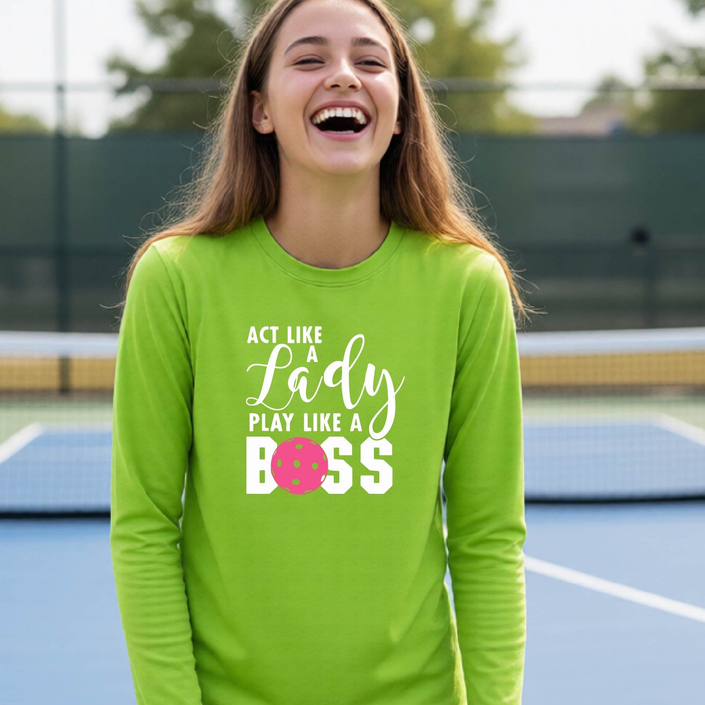 A young person laughing on a pickleball court wearing a lime green long-sleeve t-shirt with the text 'ACT LIKE A Lady PLAY LIKE A BOSS' and a pink polka-dot graphic.
