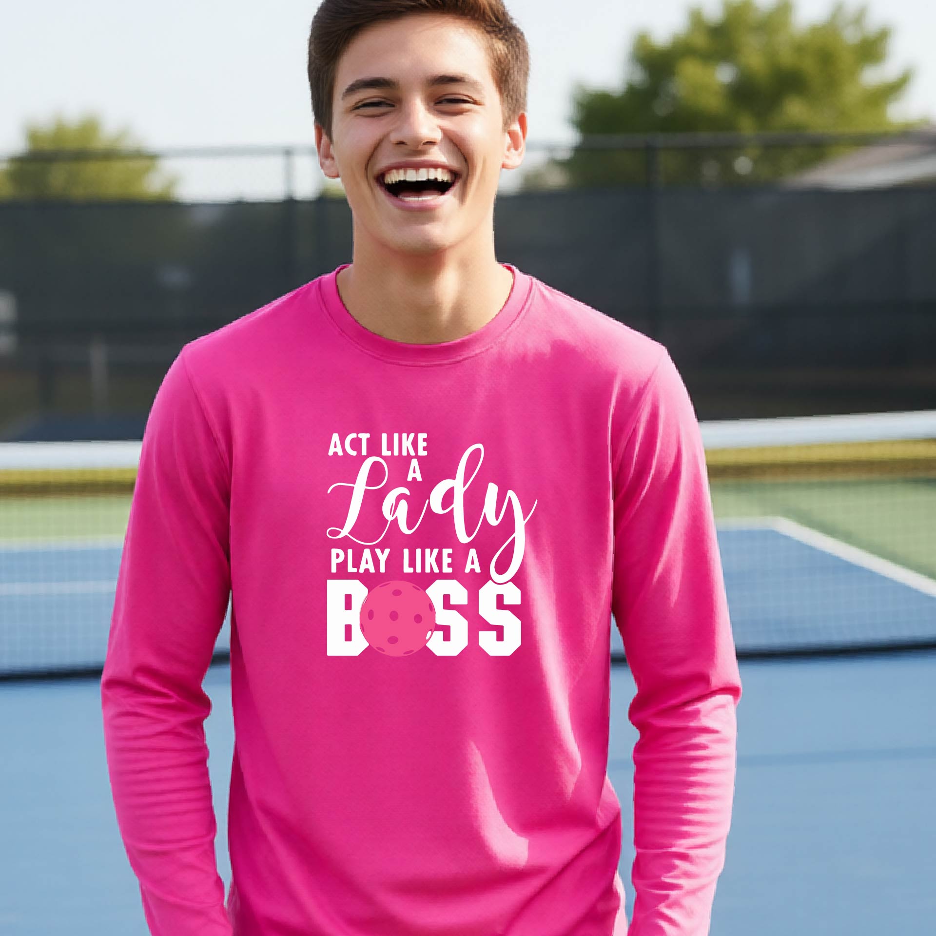 A young person laughing on a pickleball court wearing a bright pink long-sleeve t-shirt with the text 'ACT LIKE A Lady PLAY LIKE A BOSS' and a pink polka-dot graphic.