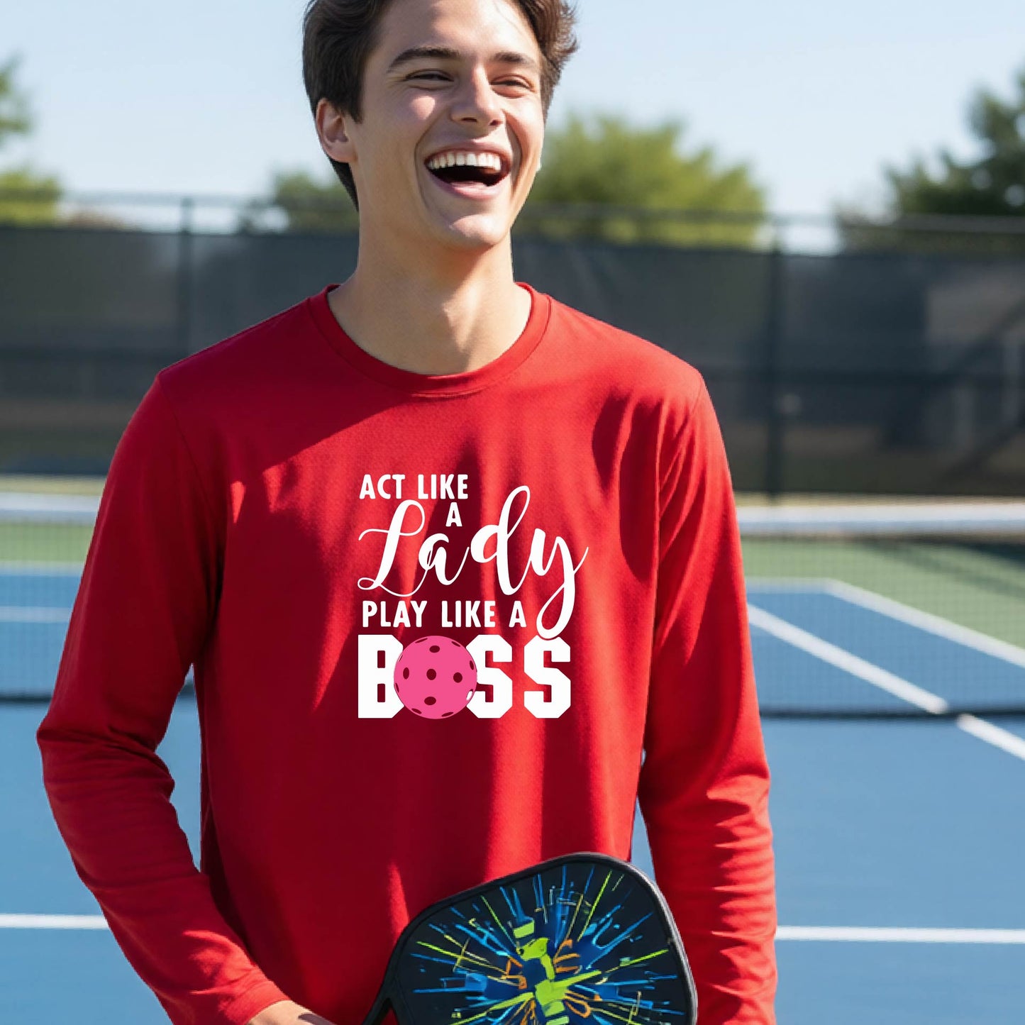 A young person laughing on a pickleball court wearing a red long-sleeve t-shirt with the text 'ACT LIKE A Lady PLAY LIKE A BOSS' and a pink polka-dot graphic.