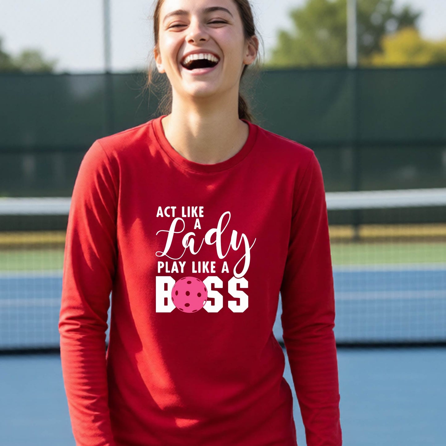 A young person laughing on a pickleball court wearing a red long-sleeve t-shirt with the text 'ACT LIKE A Lady PLAY LIKE A BOSS' and a pink polka-dot graphic.