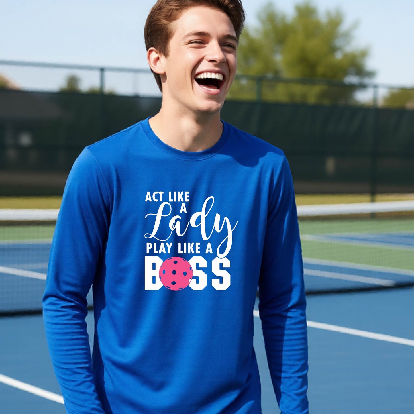 A young person laughing on a pickleball court wearing a royal blue long-sleeve t-shirt with the text 'ACT LIKE A Lady PLAY LIKE A BOSS' and a pink polka-dot graphic.