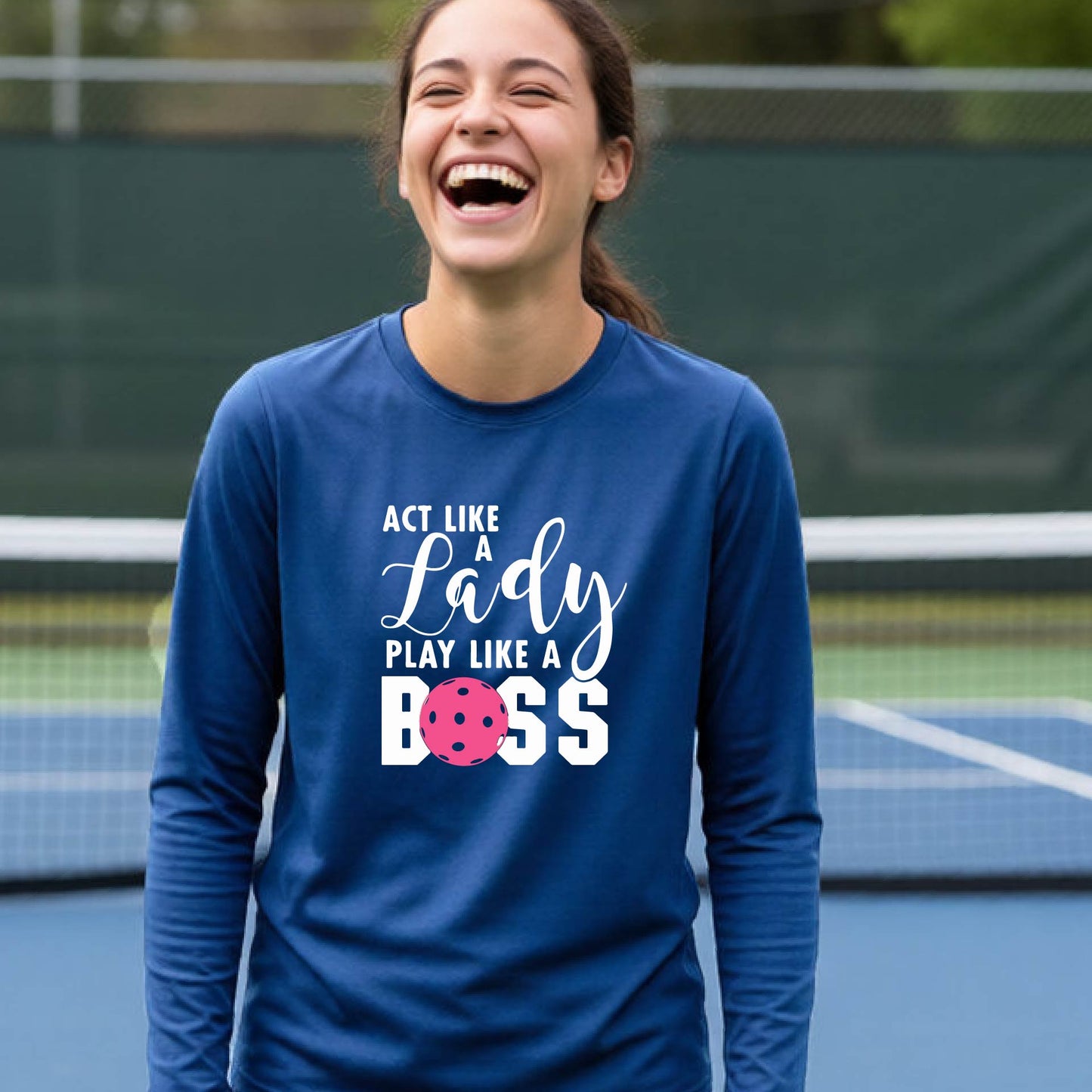 A young person laughing on a pickleball court wearing a royal blue long-sleeve t-shirt with the text 'ACT LIKE A Lady PLAY LIKE A BOSS' and a pink polka-dot graphic.