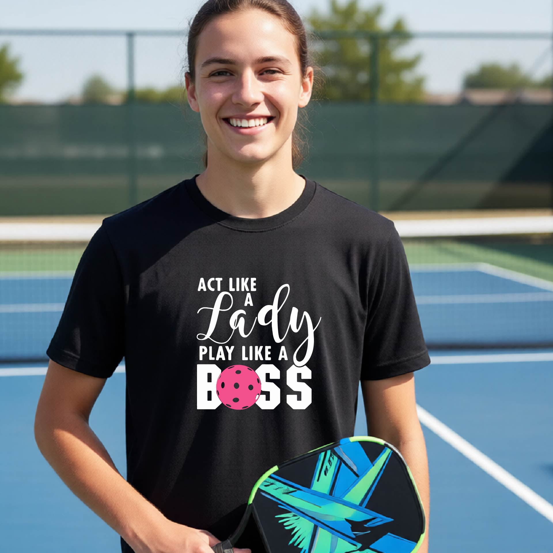 A young person laughing on an outdoor pickleball court wearing a black t-shirt with a graphic that reads 'ACT LIKE A Lady PLAY LIKE A BOSS' with a pink pickleball graphic.