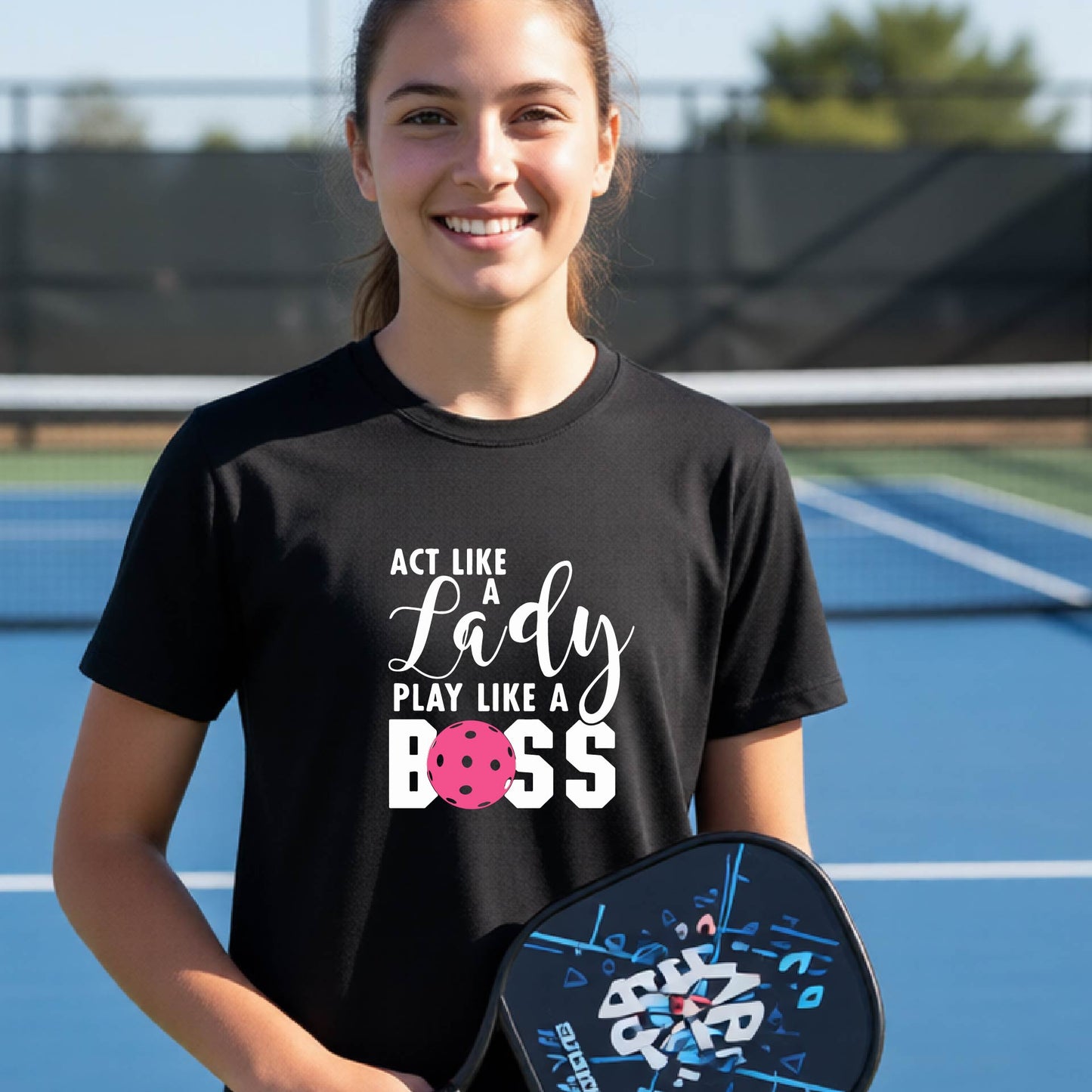 A young person laughing on an outdoor pickleball court wearing black  t-shirt with a graphic that reads 'ACT LIKE A Lady PLAY LIKE A BOSS' with a pink pickleball graphic.