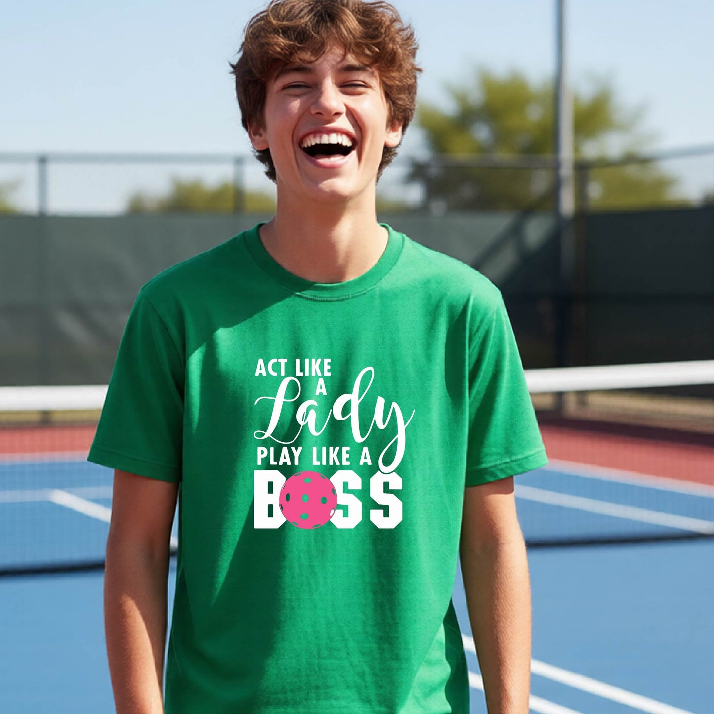 A young person laughing on an outdoor pickleball court wearing a green t-shirt with a graphic that reads 'ACT LIKE A Lady PLAY LIKE A BOSS' with a pink pickleball graphic.