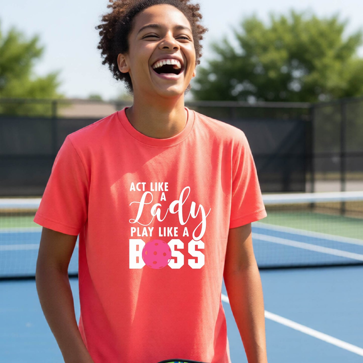 A young person laughing on an outdoor pickleball court wearing a coral pink t-shirt with a graphic that reads 'ACT LIKE A Lady PLAY LIKE A BOSS' with a pink pickleball graphic.