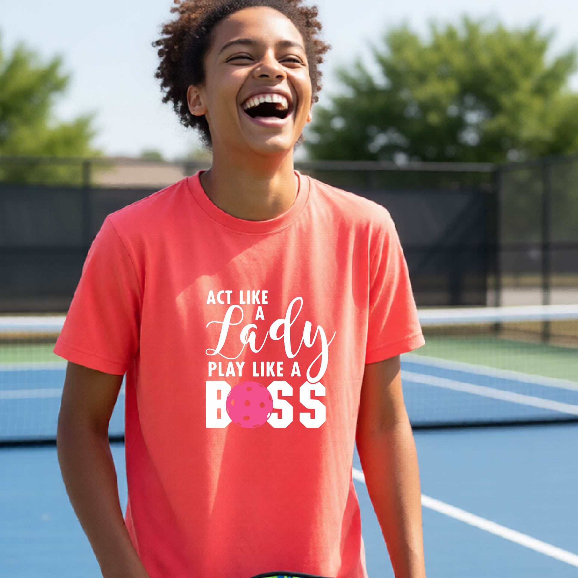 A young person laughing on an outdoor pickleball court wearing a coral pink t-shirt with a graphic that reads 'ACT LIKE A Lady PLAY LIKE A BOSS' with a pink pickleball graphic.