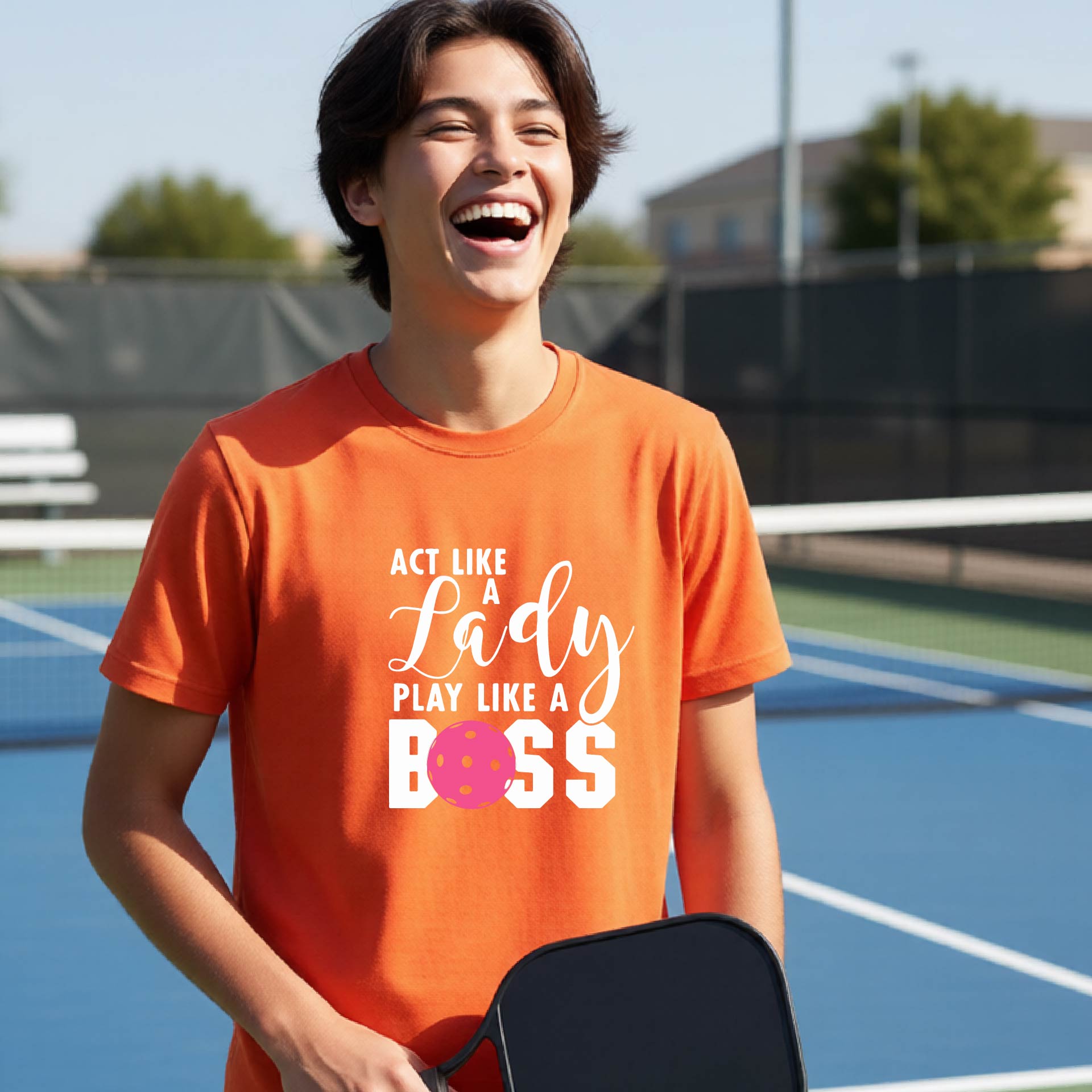 A young person laughing on an outdoor pickleball court wearing an orange t-shirt with a graphic that reads 'ACT LIKE A Lady PLAY LIKE A BOSS' with a pink pickleball graphic.