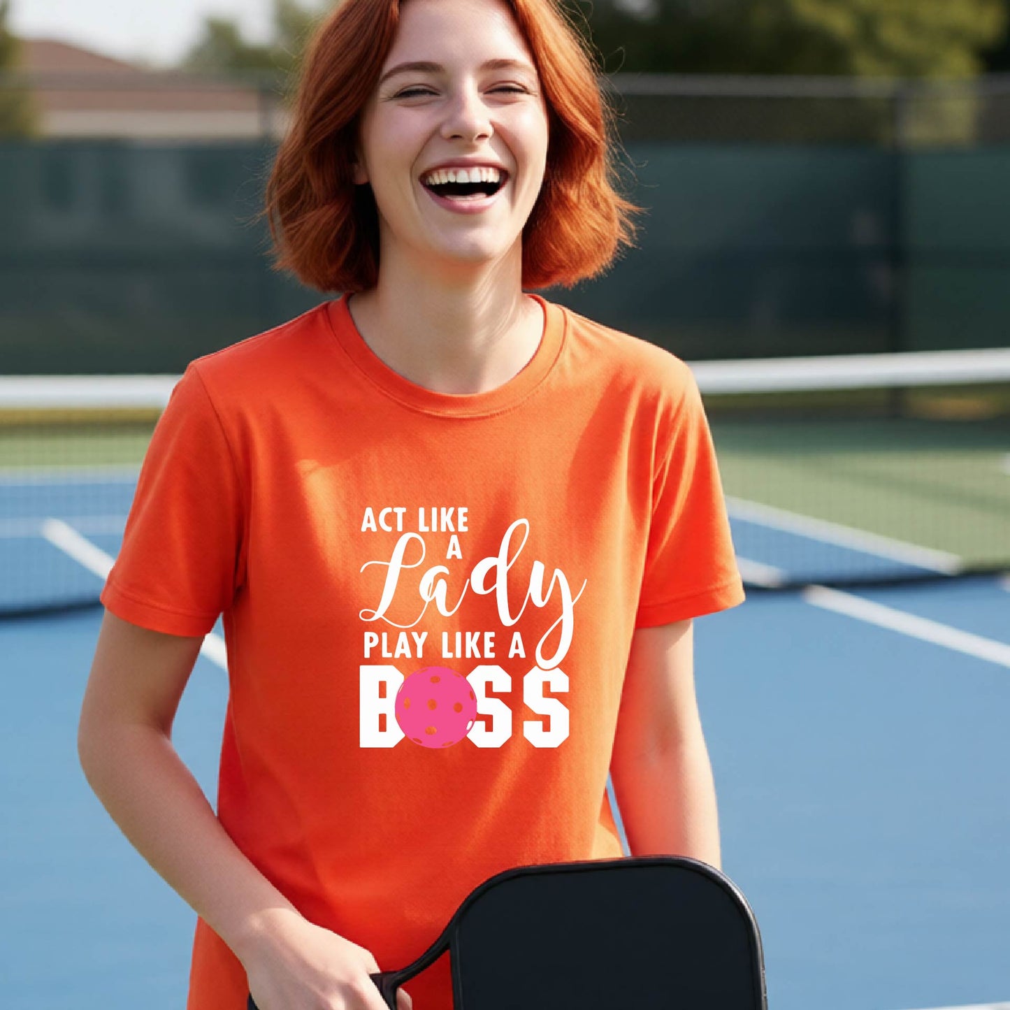A young person laughing on an outdoor pickleball court wearing an orange t-shirt with a graphic that reads 'ACT LIKE A Lady PLAY LIKE A BOSS' with a pink pickleball graphic.