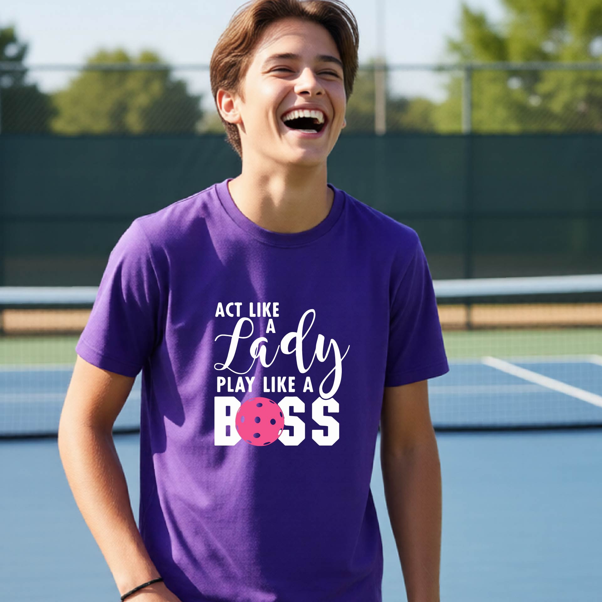 A young person laughing on an outdoor pickleball court wearing a purple t-shirt with a graphic that reads 'ACT LIKE A Lady PLAY LIKE A BOSS' with a pink pickleball graphic.